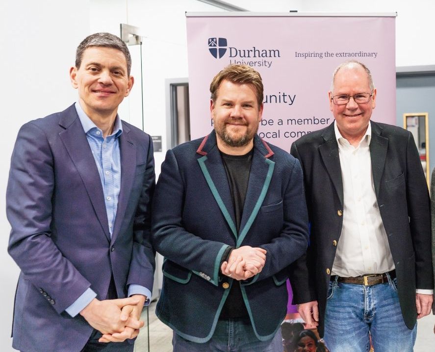 Four men, including James Corden and Joe Docherty, standing in front of a Durham University banner.