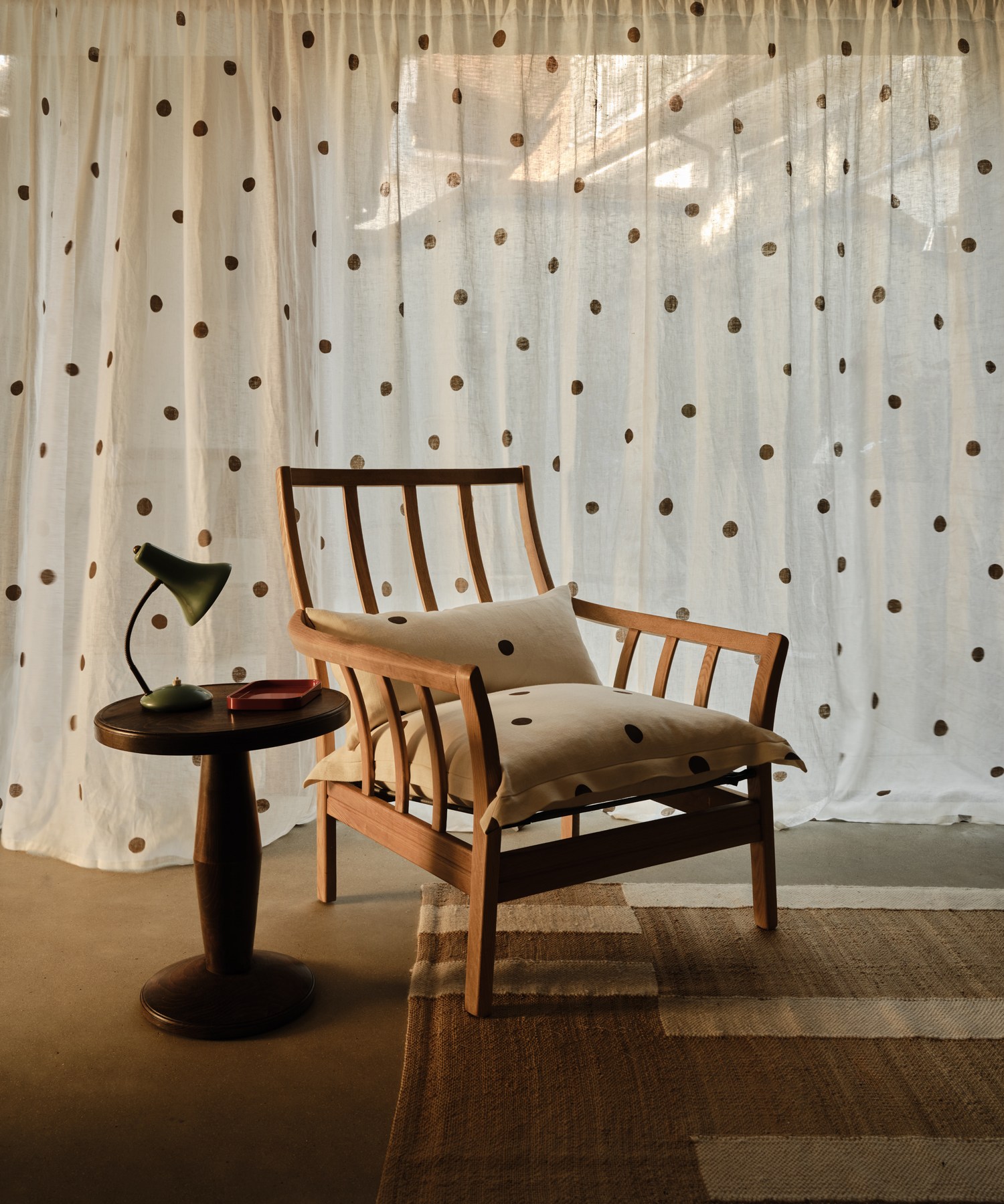 A wooden armchair with spotted cushions beside a round side table with a lamp, in front of a white curtain with brown polka dots.