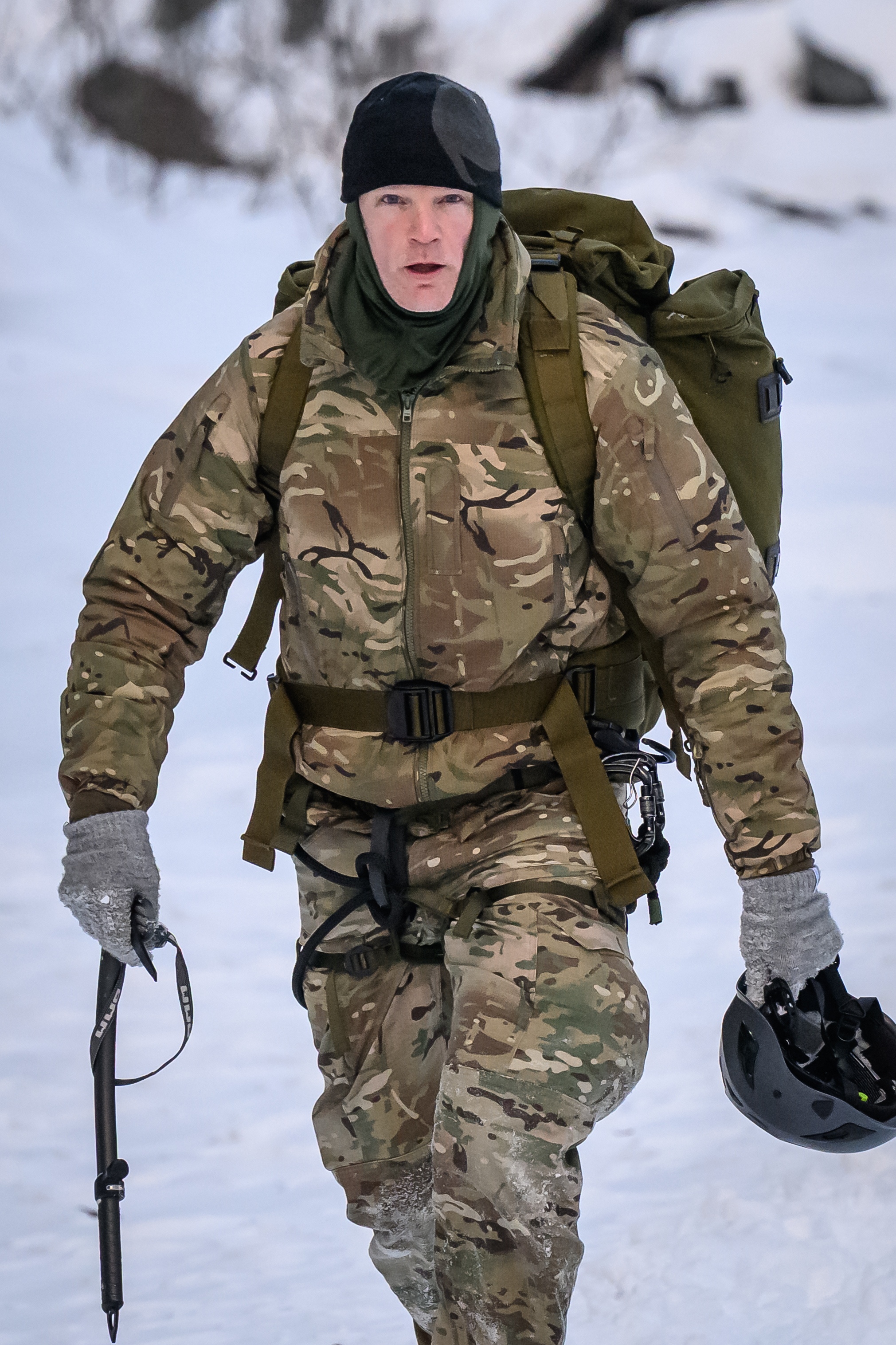 UK Armed Forces Minister Al Carns in camouflage, wearing a black beanie and neck gaiter, a backpack, and carrying an ice axe and helmet in a snowy landscape.