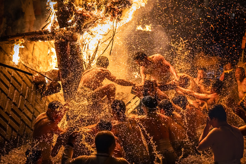 Men in a Japanese festival carrying a burning structure while being splashed with water.