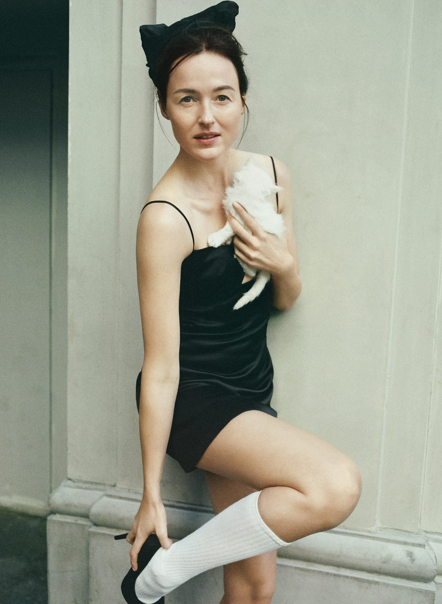 Renate Reinsve wearing a black slip dress, black kitten-ear headband, white knee-high socks, and black heels, holding a white fluffy puppy.