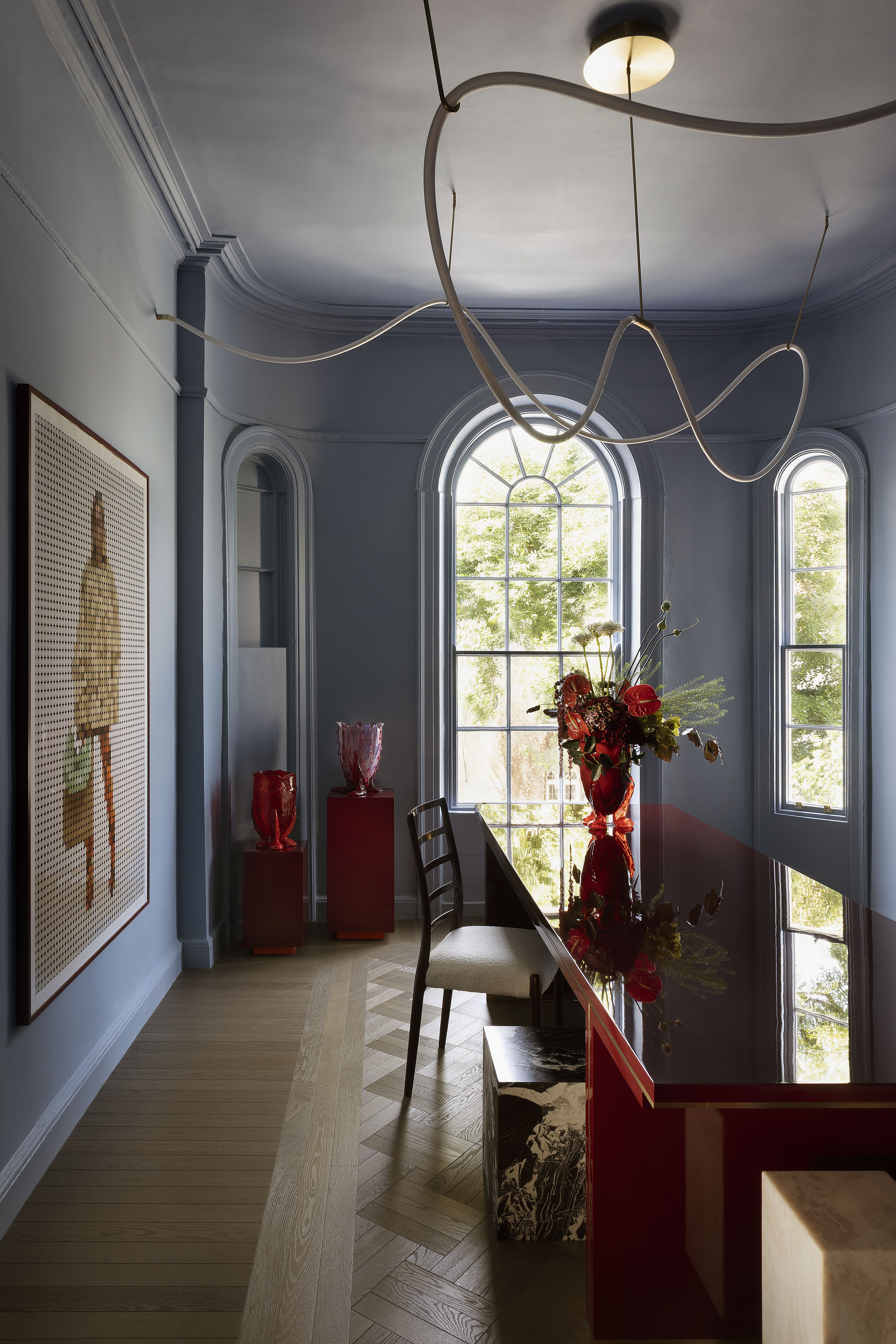 Dining room with light blue walls, two arched windows, a red glossy table, a decorative light fixture, and abstract artwork.