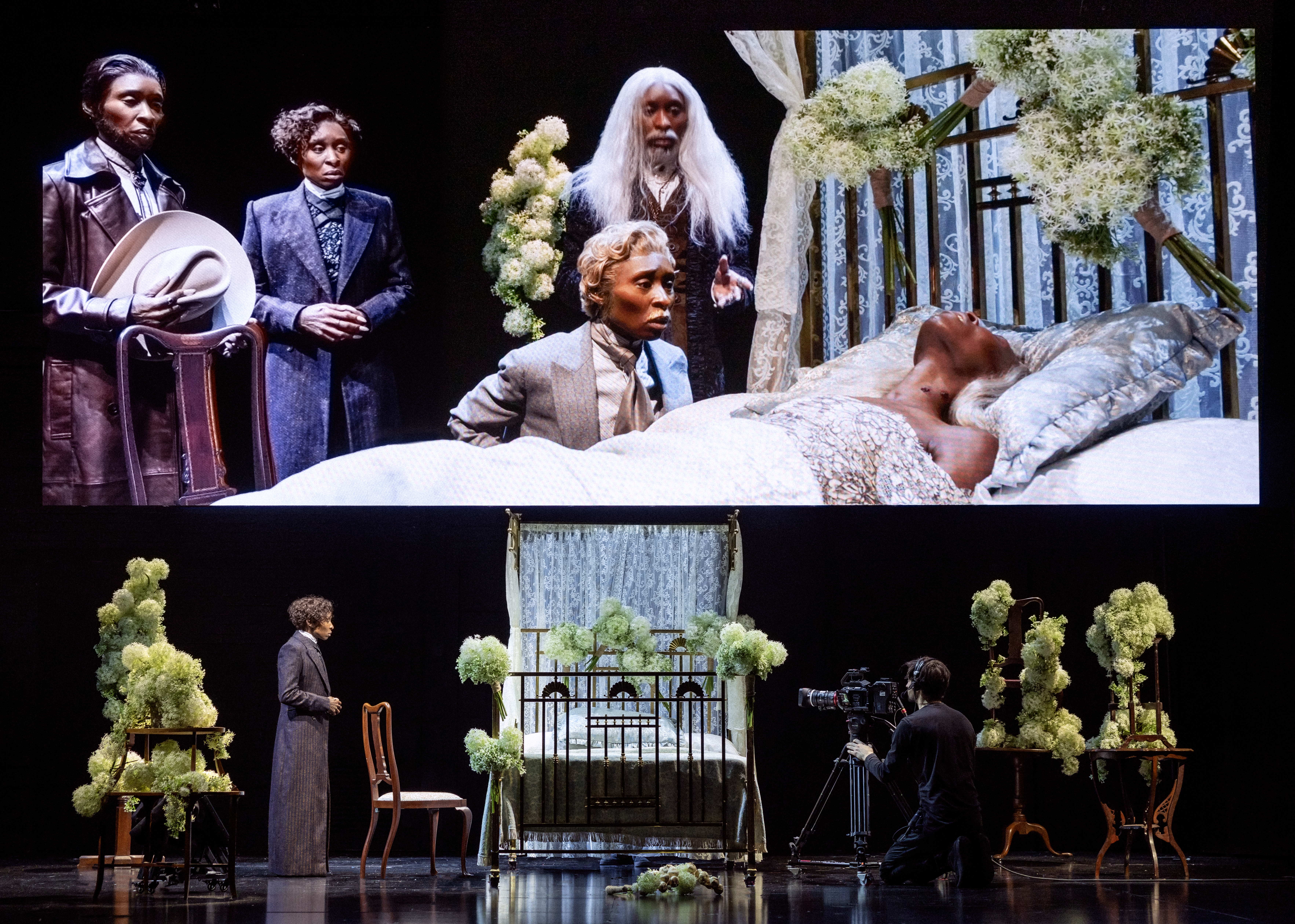 Cynthia Erivo and other actors perform in a scene from Dracula, with a projection behind them showing close-ups of the actors on a bed.