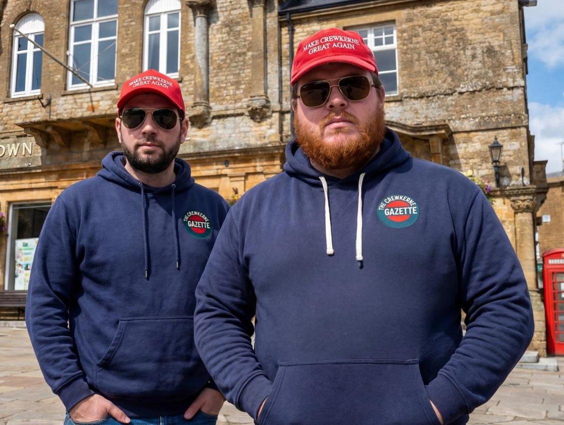 Two men in "Make Crewkerne Great Again" hats and "The Crewkerne Gazette" hoodies stand in front of a building.