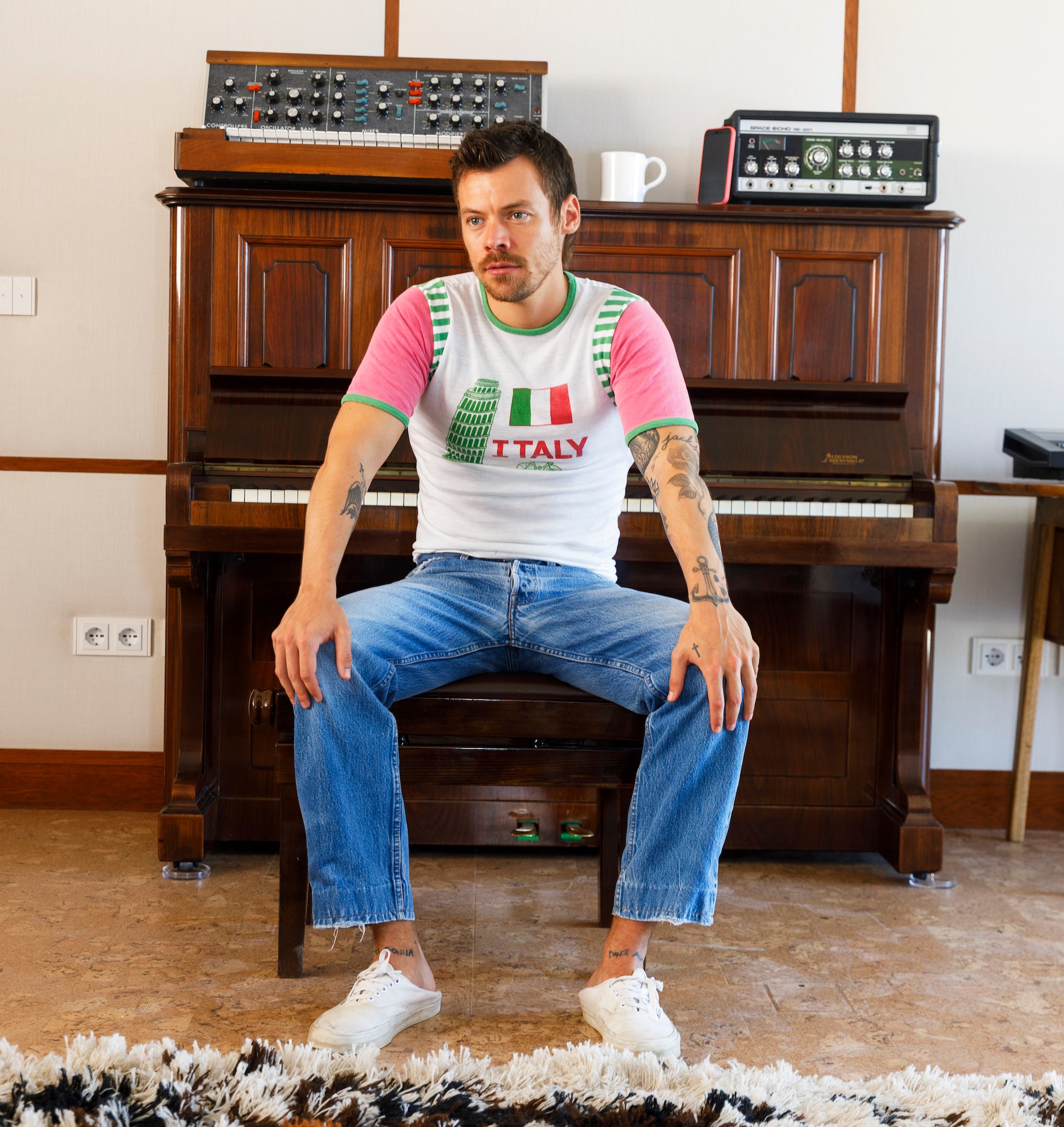 Harry Styles sitting on a piano bench wearing a white t-shirt with an Italian flag and "ITALY" printed on it.