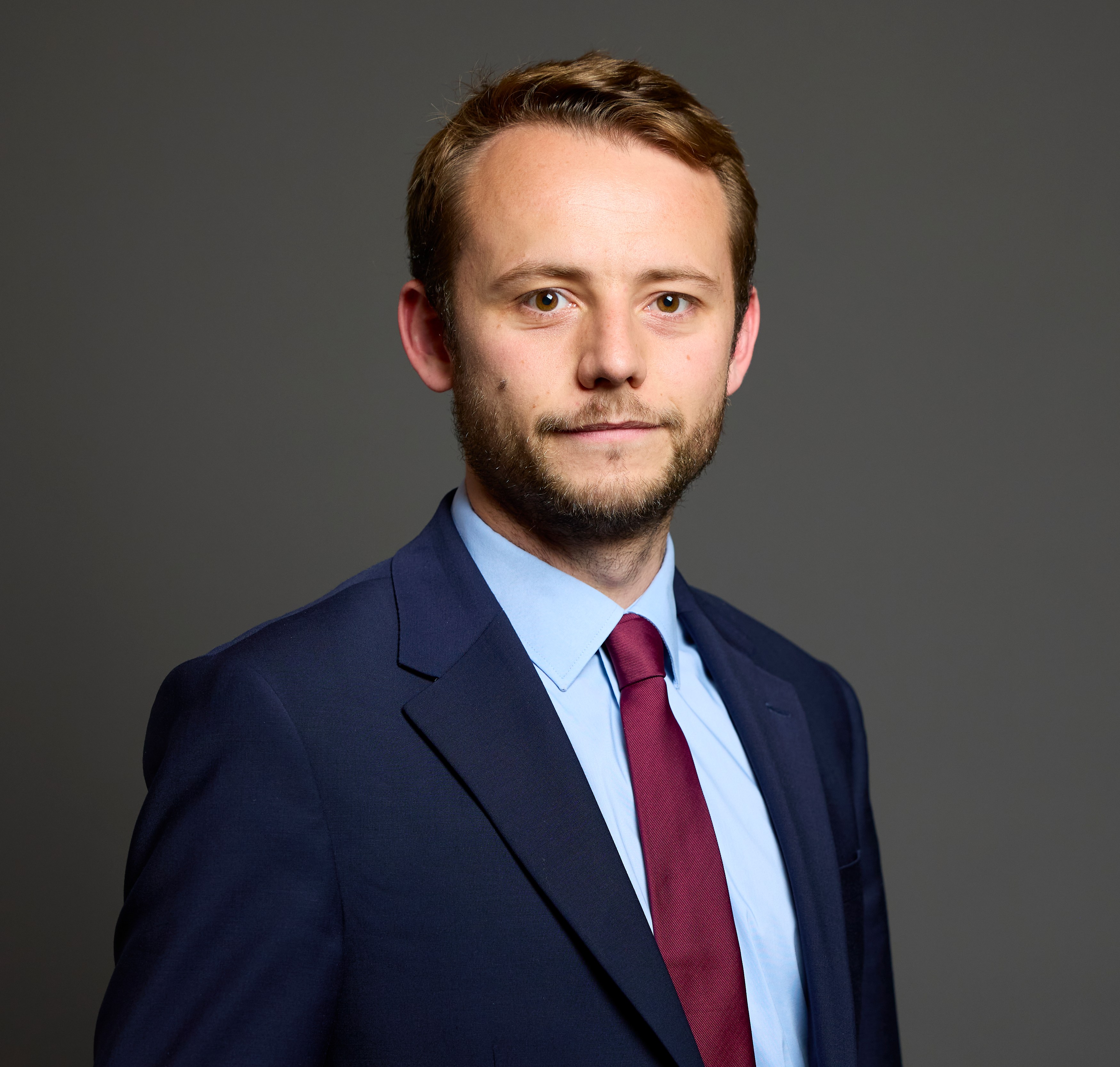 A man in a navy suit, light blue shirt, and red tie stands facing forward.
