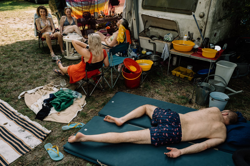 A group of people relaxing outdoors, some sitting in chairs while one man lies on a mat, next to a makeshift outdoor kitchen area with a camper van.