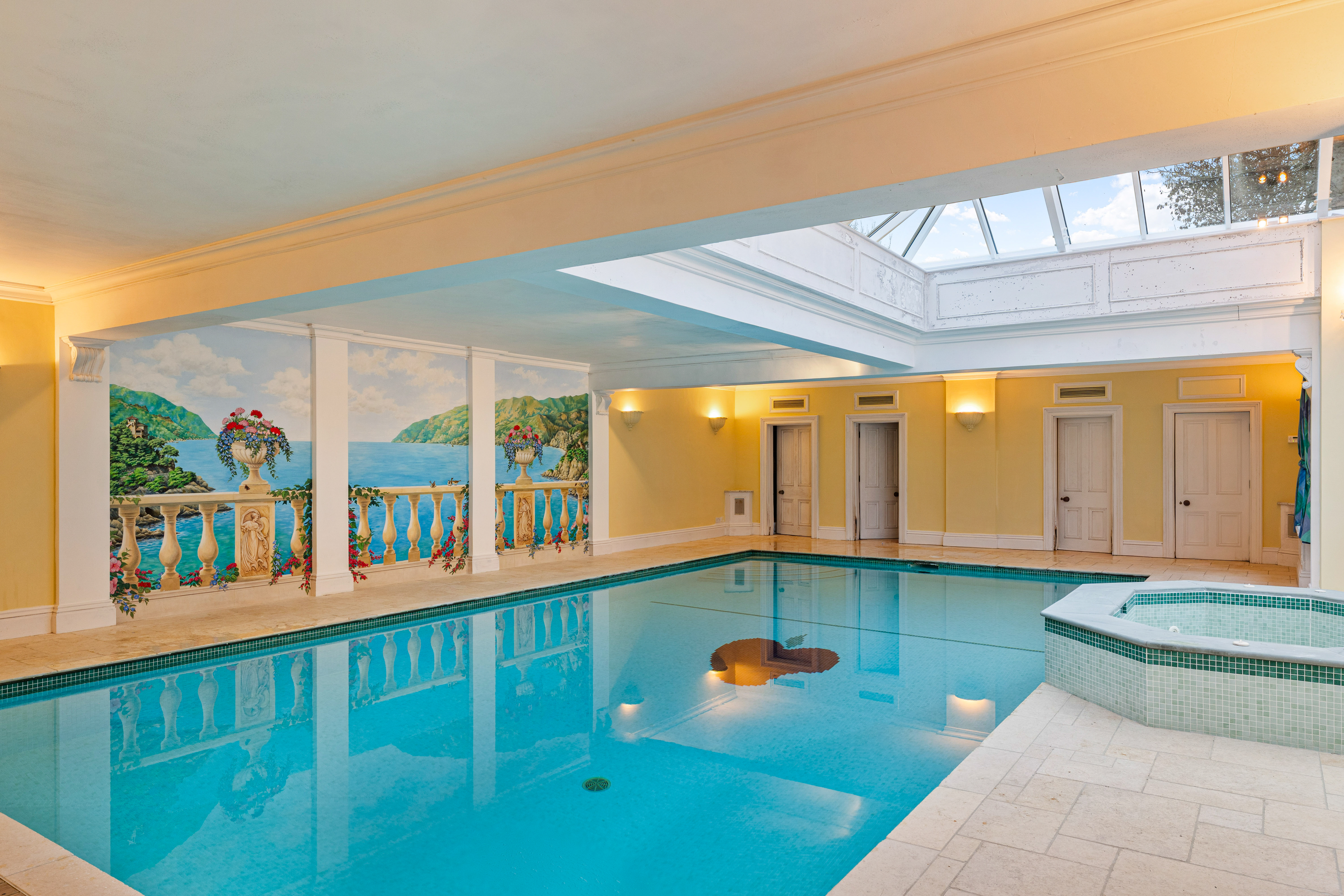 Indoor swimming pool with an adjacent hot tub, a skylight, and a mural of a seaside landscape.