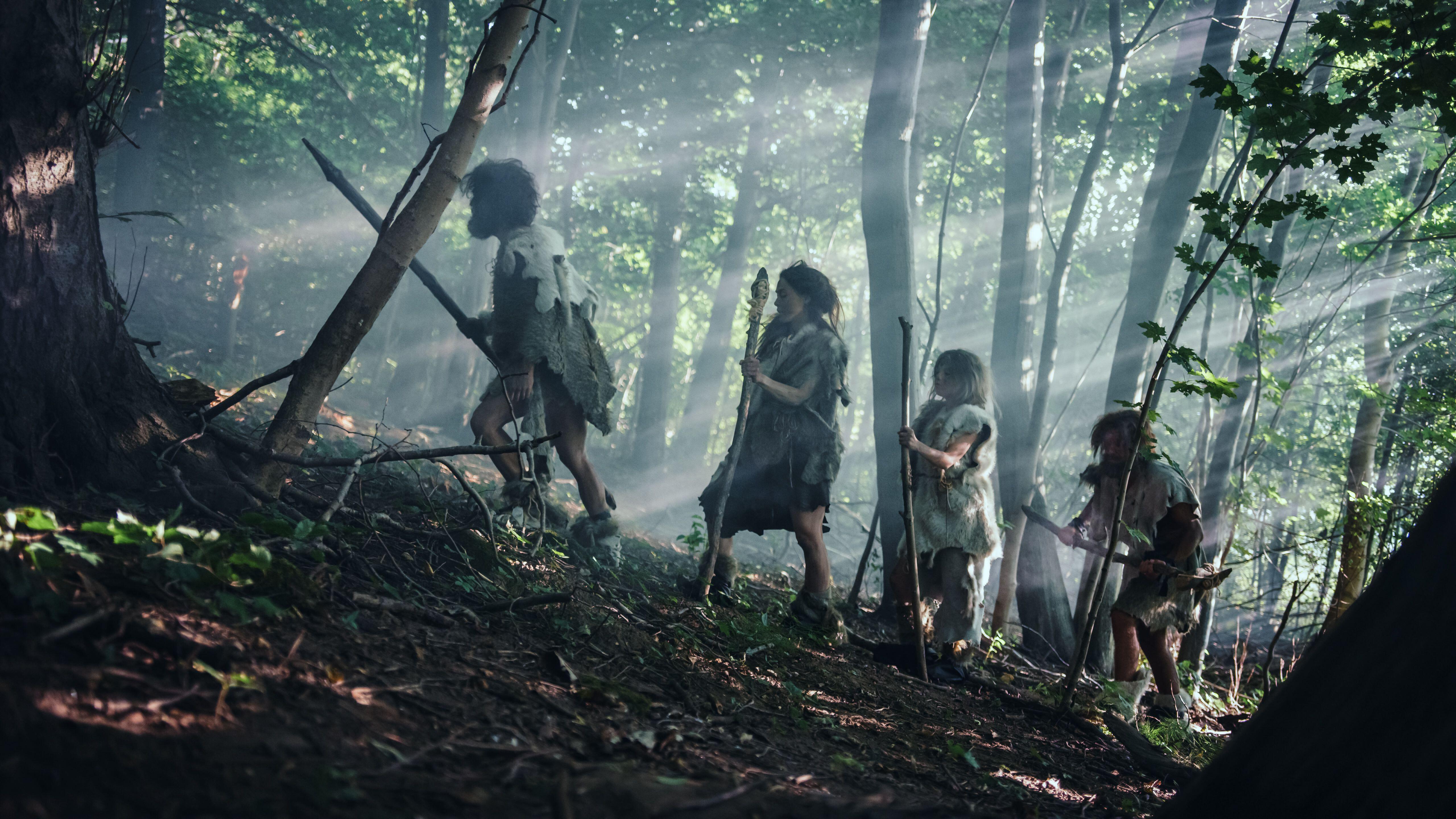 A Neanderthal family, dressed in animal skins and carrying stone-tipped tools, walks through a misty, sun-dappled forest.