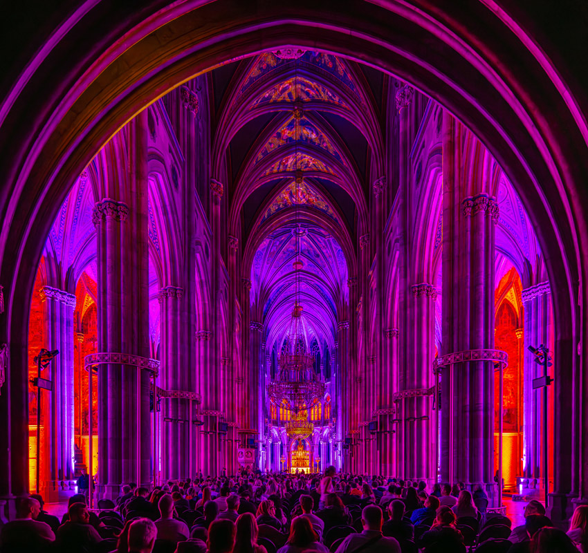 An audience is seated in a large, ornate hall with high arched ceilings, illuminated in vibrant purple and red lights.