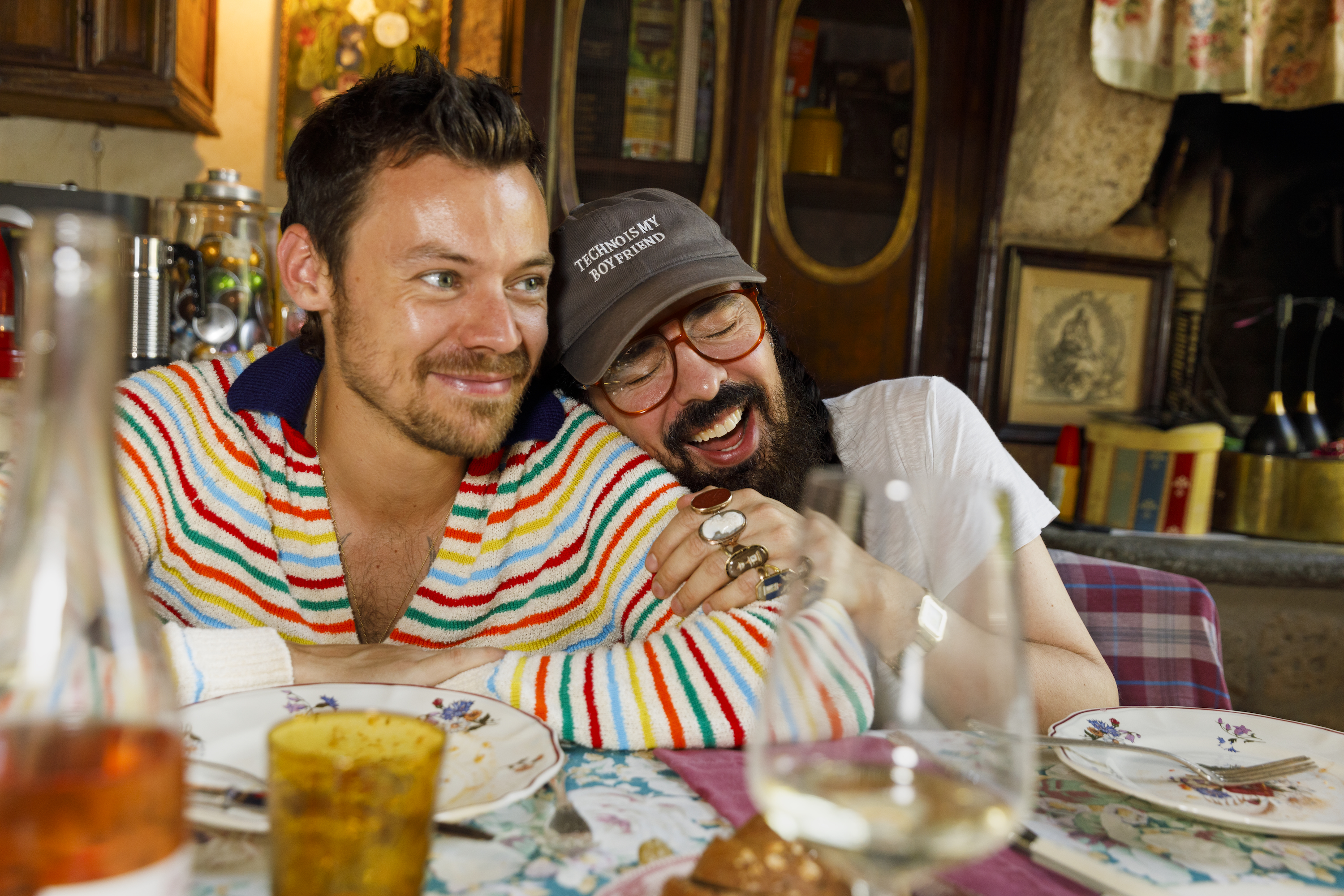 Harry Styles in a rainbow-striped sweater and a man in a "Techno is my boyfriend" hat embracing at a dining table.