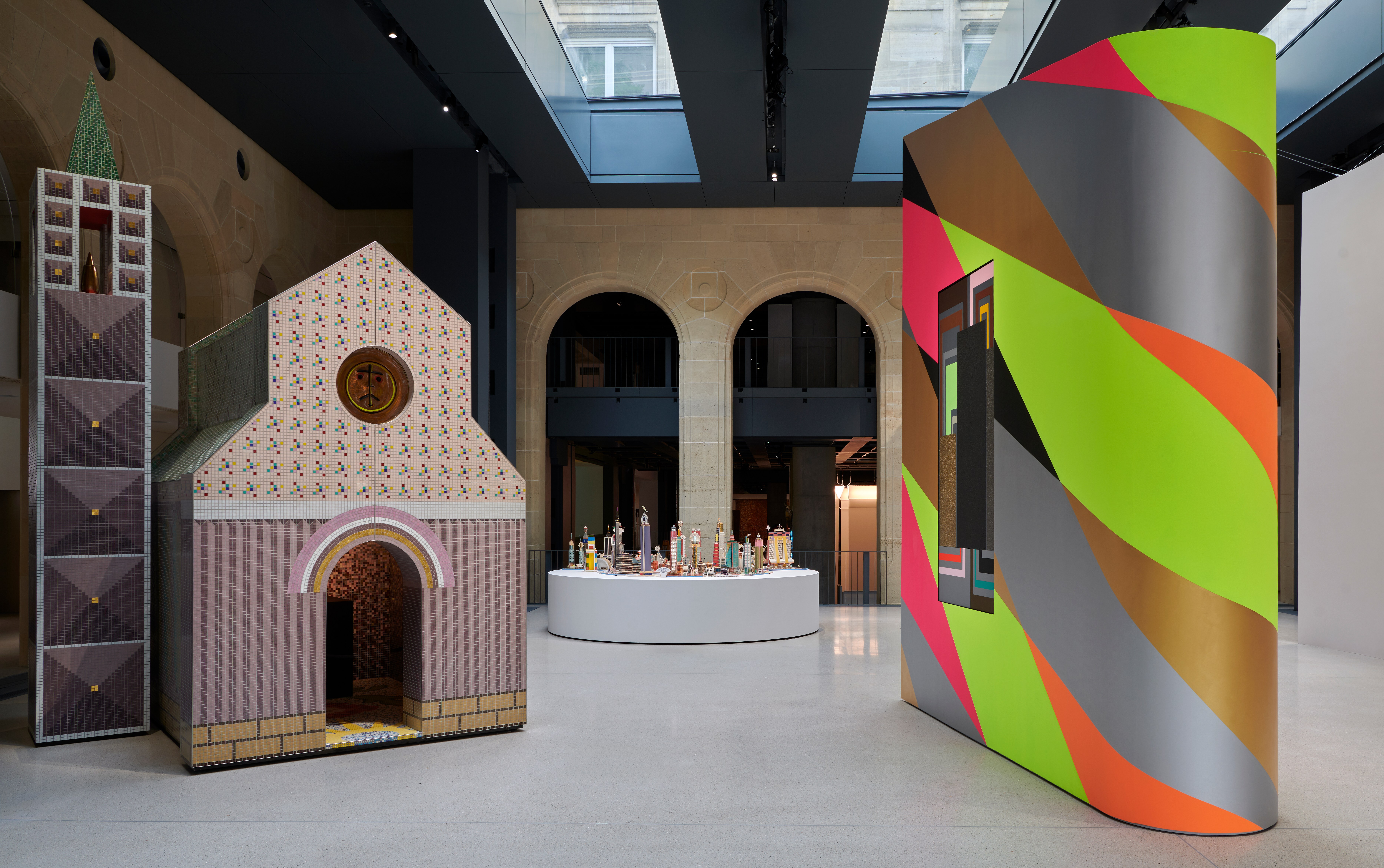 Art installation at Fondation Cartier featuring a miniature tiled building, a colorful striped wall, and a collection of small sculptures.