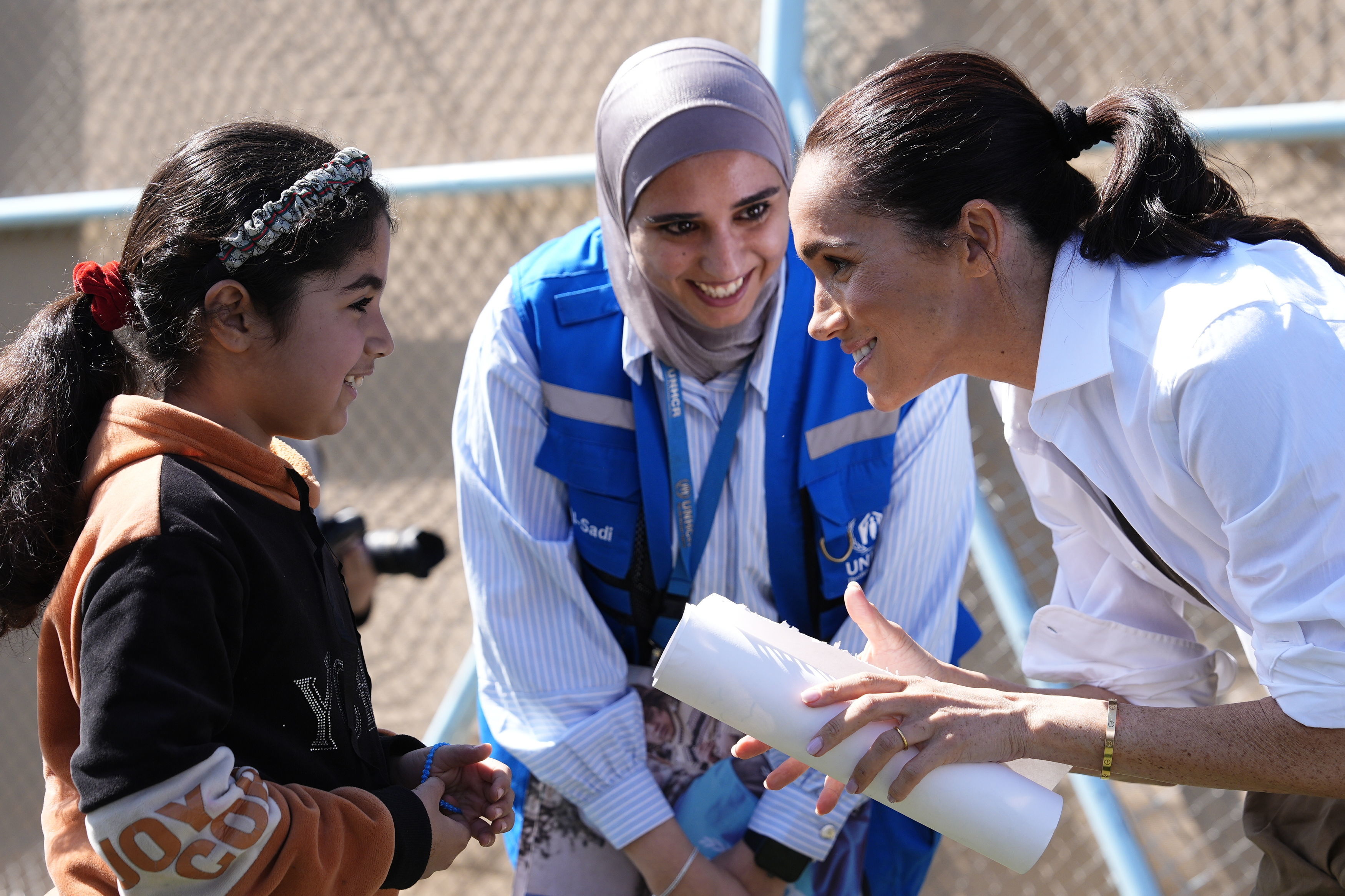 Meghan Markle speaking with a young girl at the QuestScope Youth Center in Za'atari refugee camp, while a UNHCR worker smiles in the background.