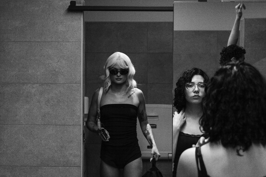 A person in sunglasses and a wig stands before a mirror reflecting a woman in glasses and another person with their arm raised.