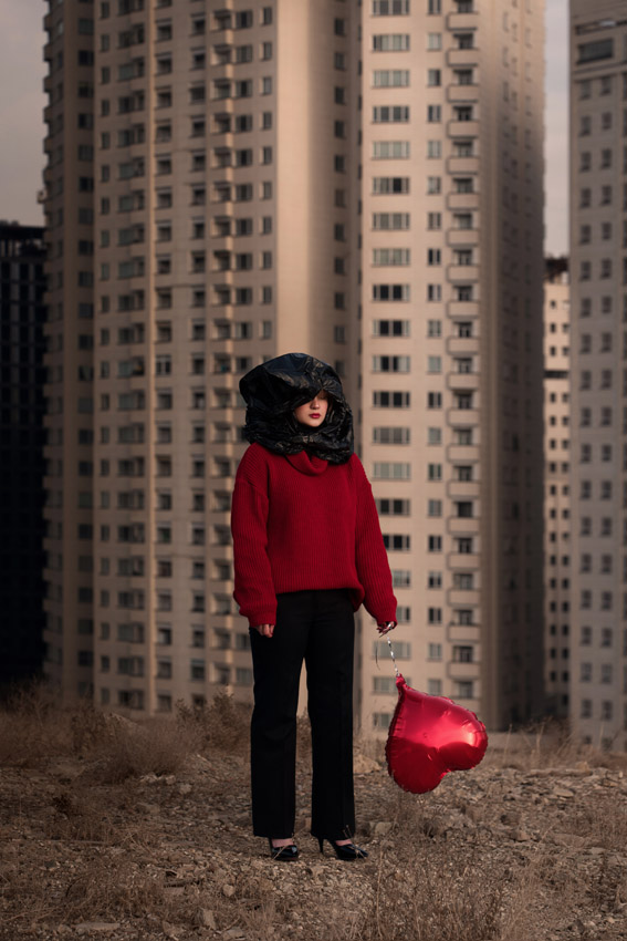 A woman with a plastic bag over her head holds a red heart-shaped balloon in front of tall buildings.