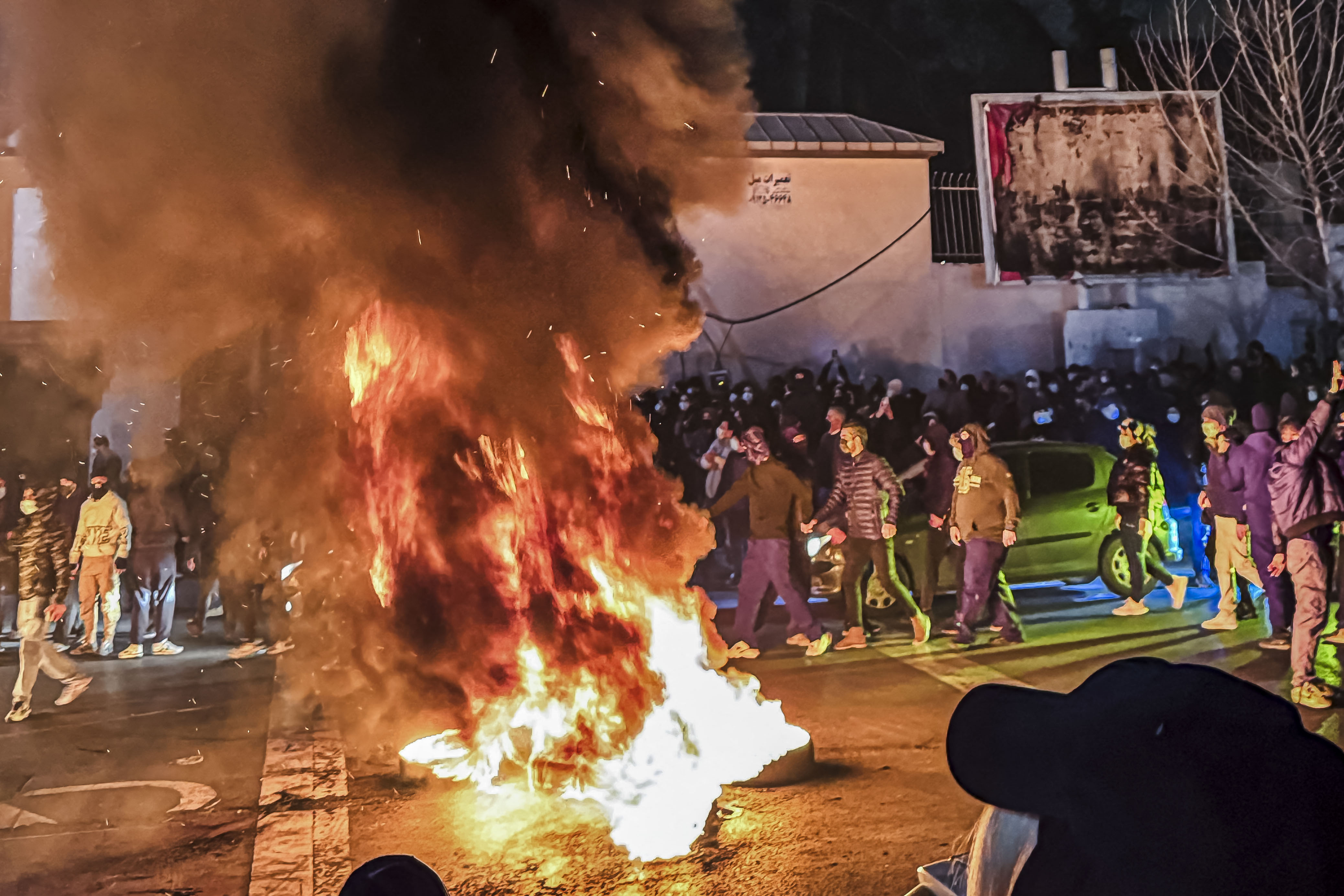 Protesters blocking a street during a demonstration in Tehran, with a large fire burning in the foreground.