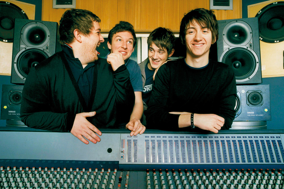 The Arctic Monkeys posing at a large audio mixing console in a studio.