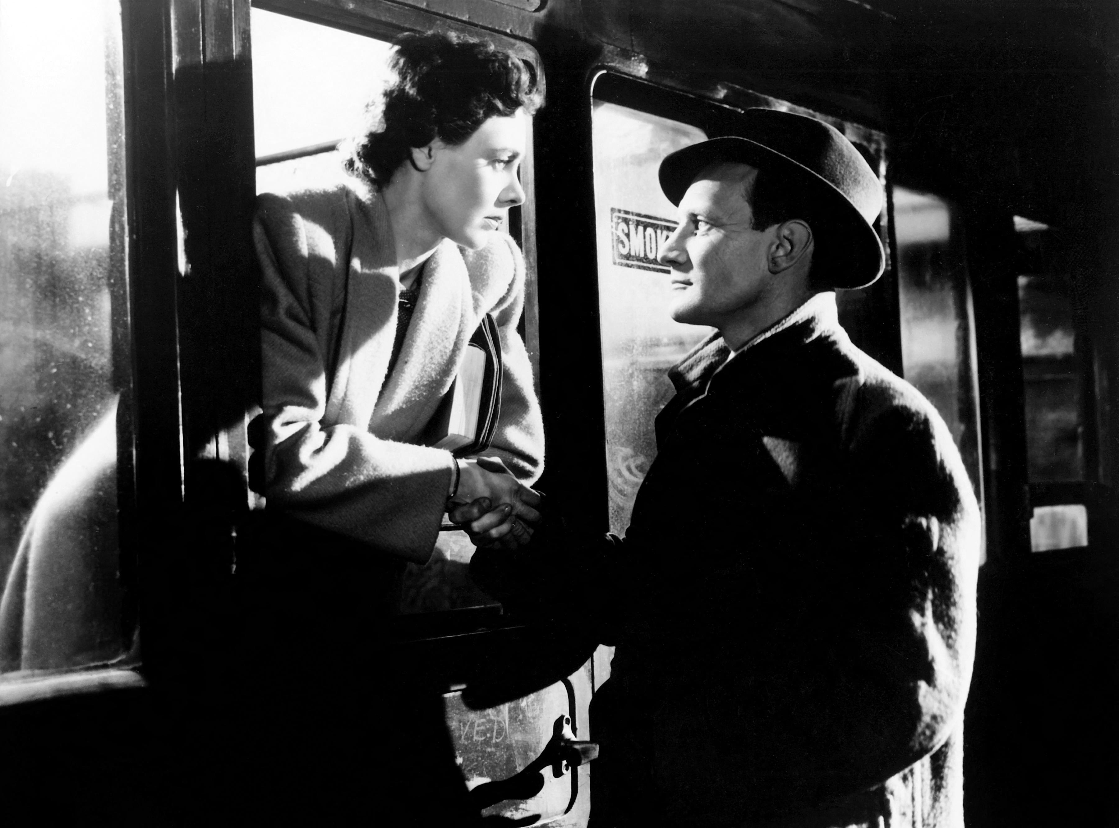 Black and white photo of a woman in a trench coat leaning out of a train window to hold hands with a man wearing a fedora.