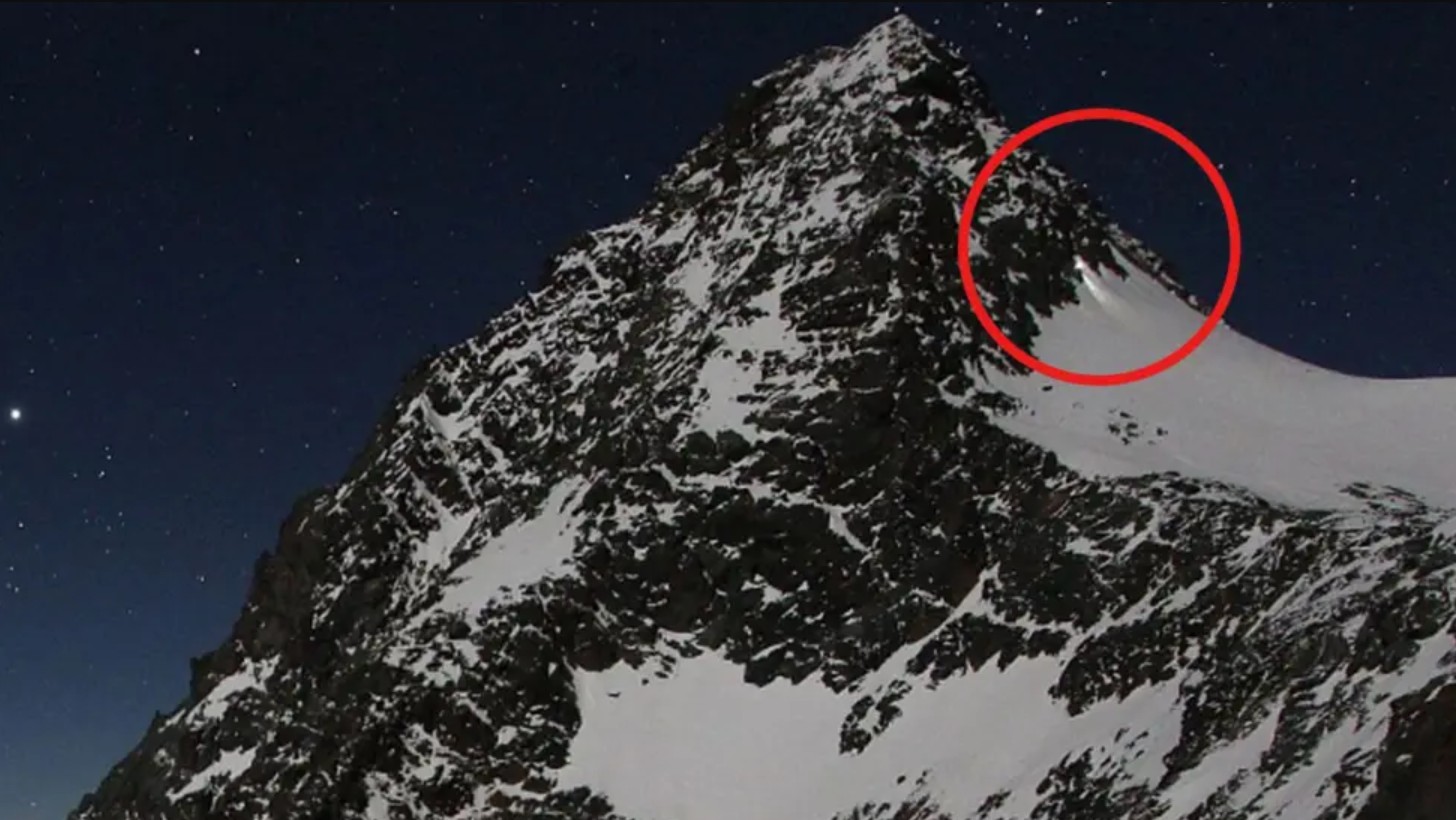 Webcam footage showing a person with a torch descending a snowy mountain peak at night.