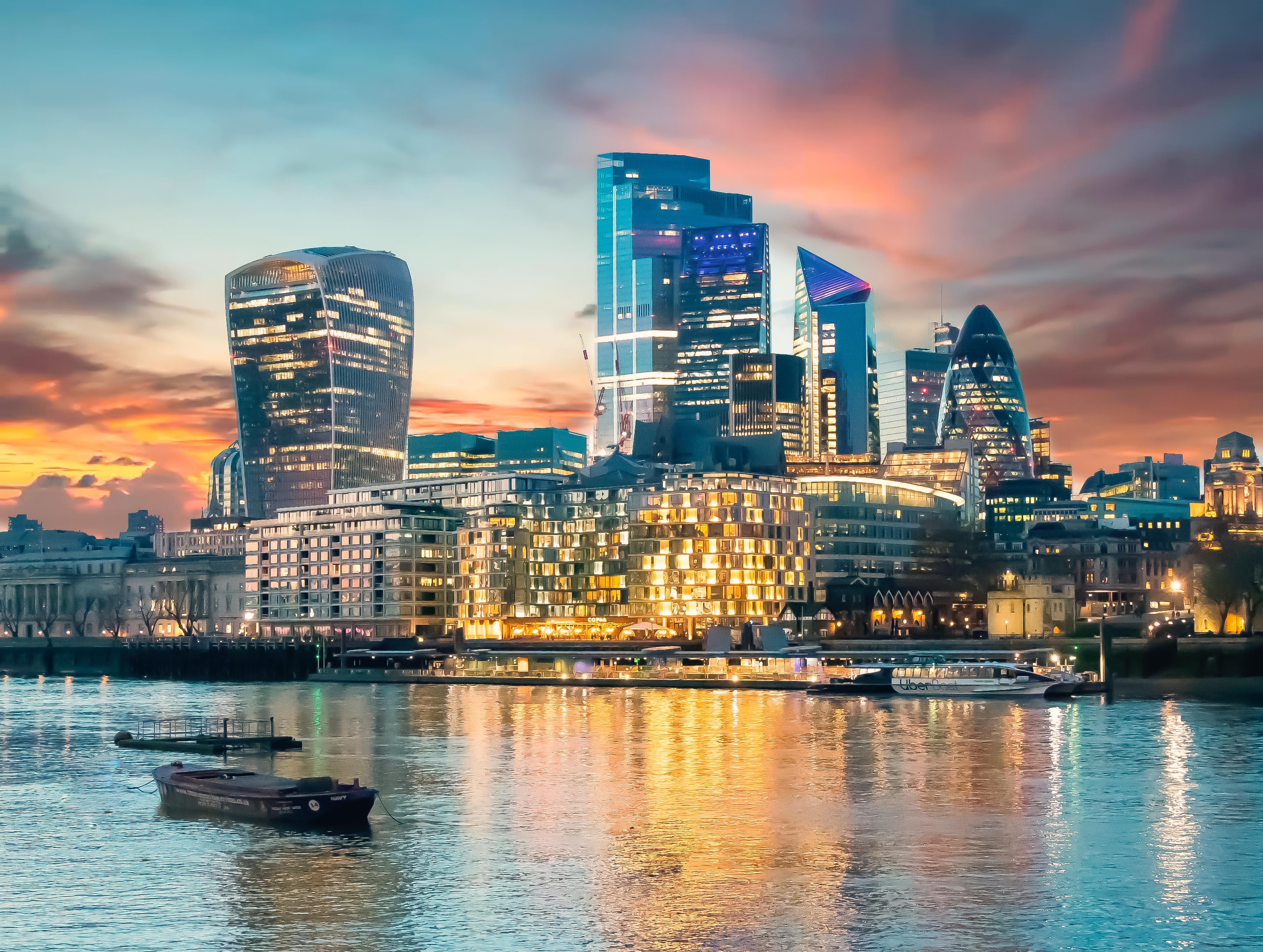 The London cityscape at dusk with the Thames River in the foreground reflecting the illuminated buildings and a colorful sky.