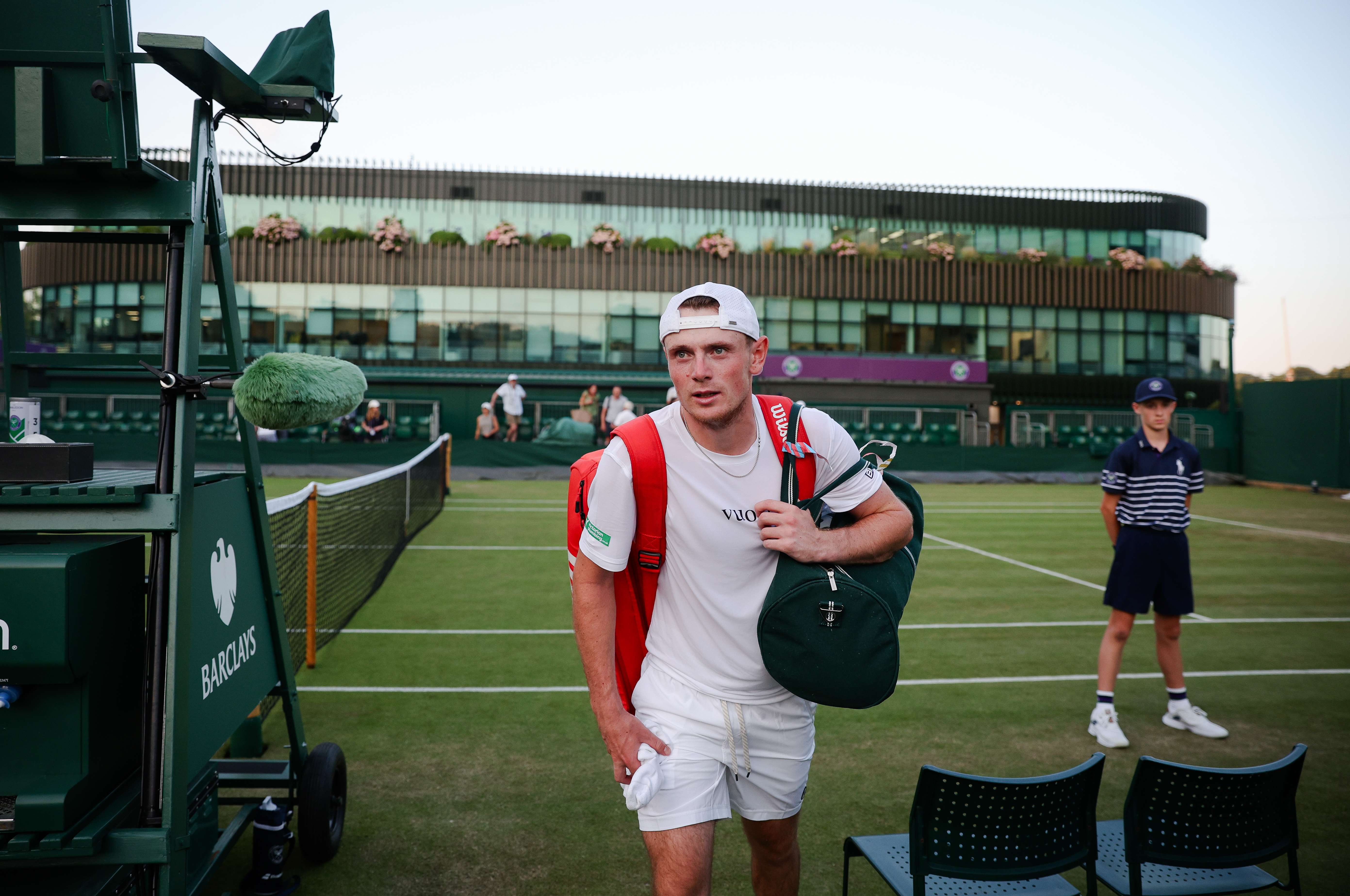 Jack Pinnington Jones, wearing a white cap, t-shirt, and shorts, stands on the tennis court with a red bag on his back and a dark green duffel bag on his shoulder.