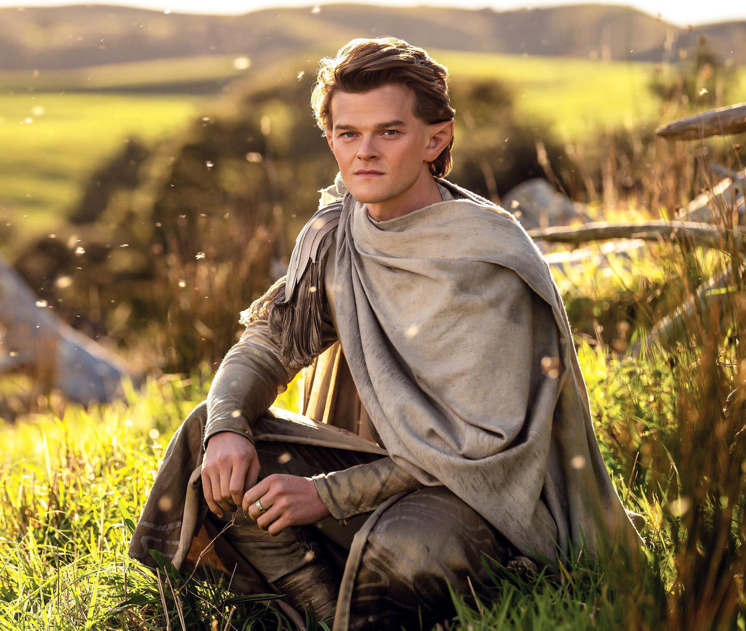 Robert Aramayo as Elrond in The Lord of the Rings: The Rings of Power, crouching in tall grass in a field.