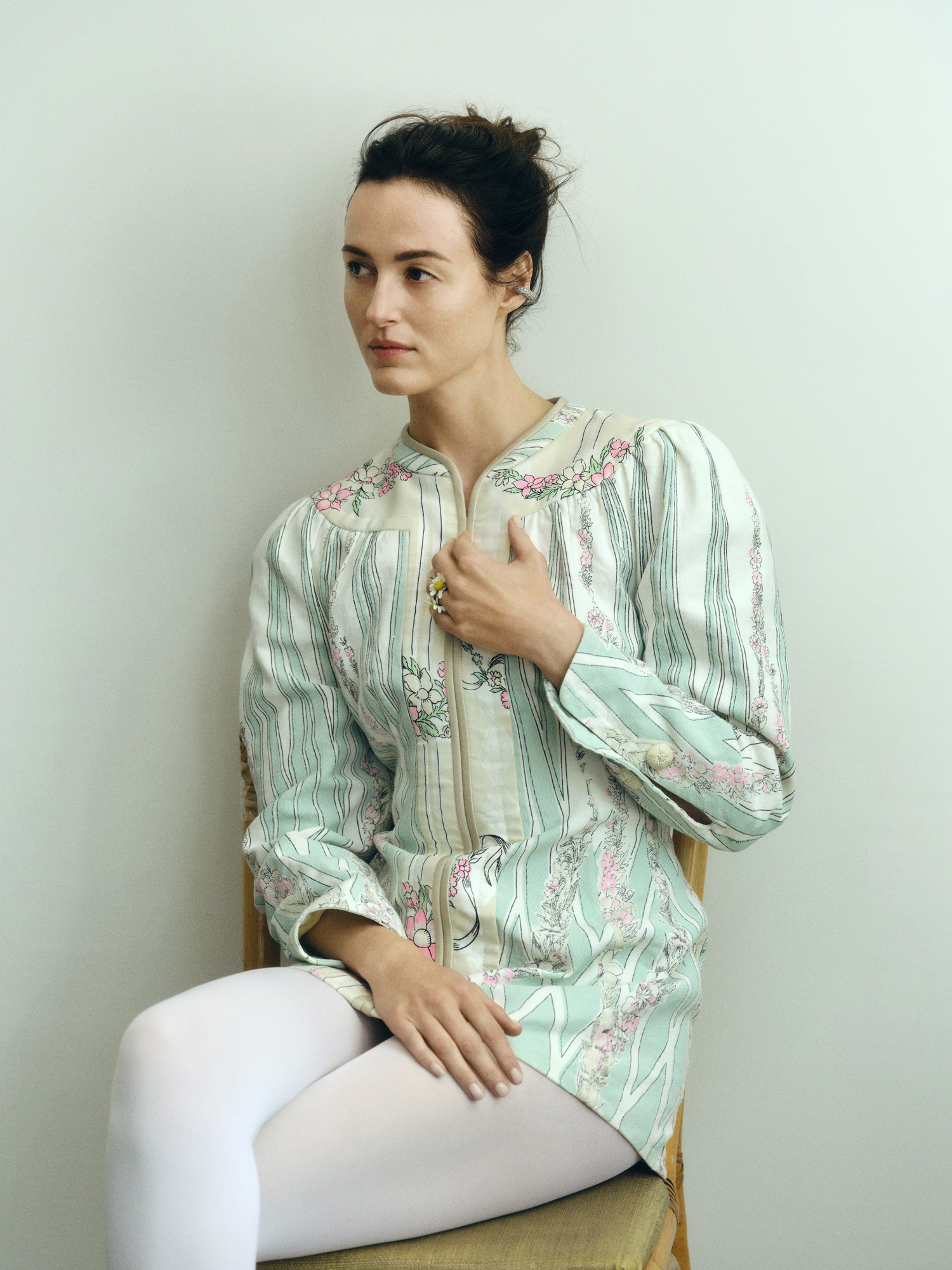 Renate Reinsve wearing a patterned jacket and white leggings, sitting against a white wall.