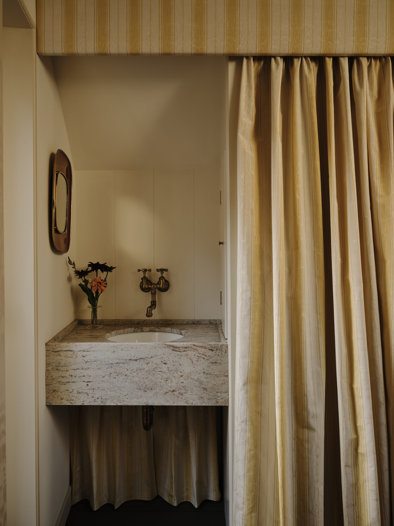 A small bathroom with a granite counter and sink, brass faucet, a mirror, and striped curtains.