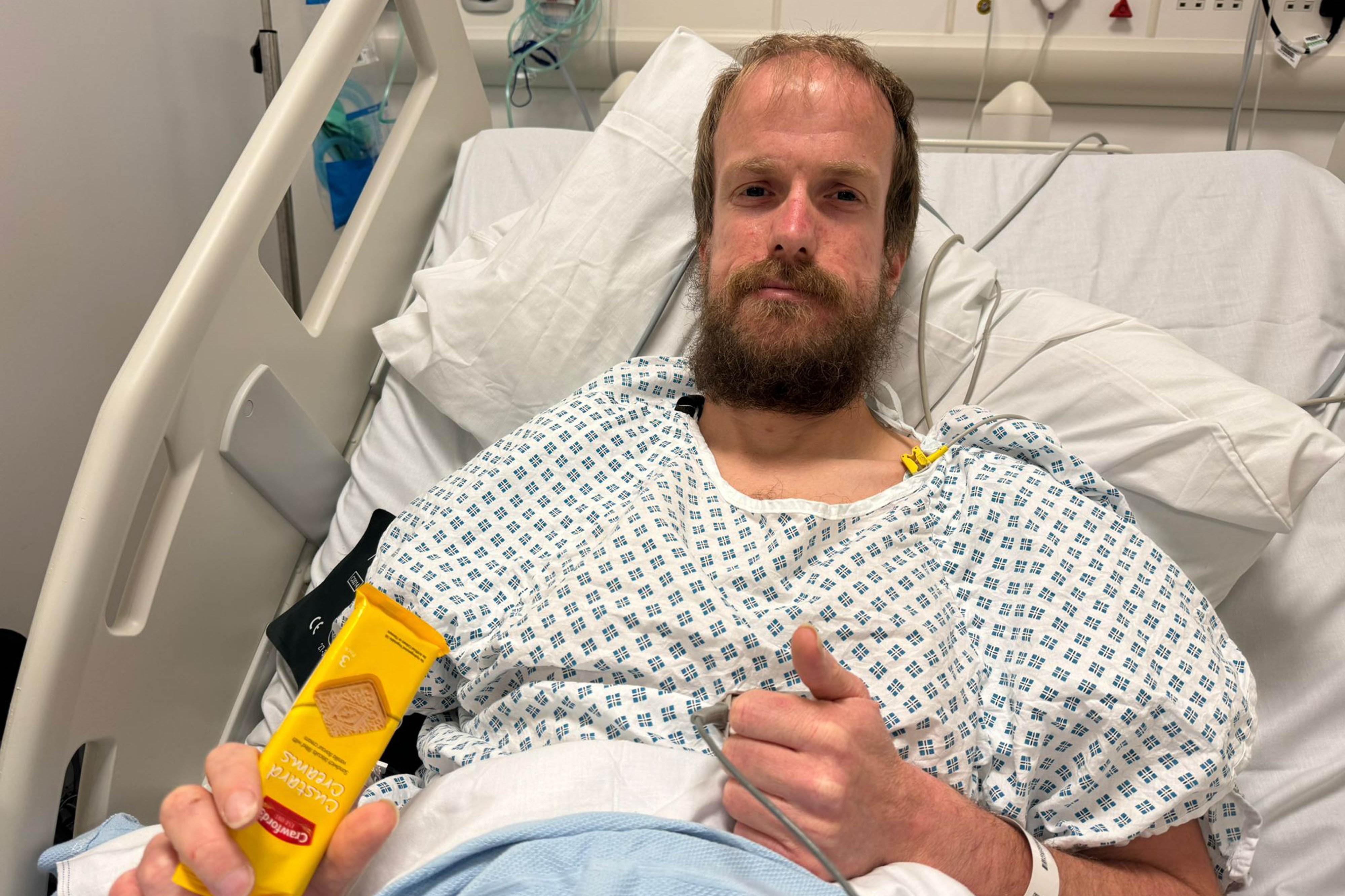 Nathaniel Dye in a hospital gown holding a packet of Custard Creams.