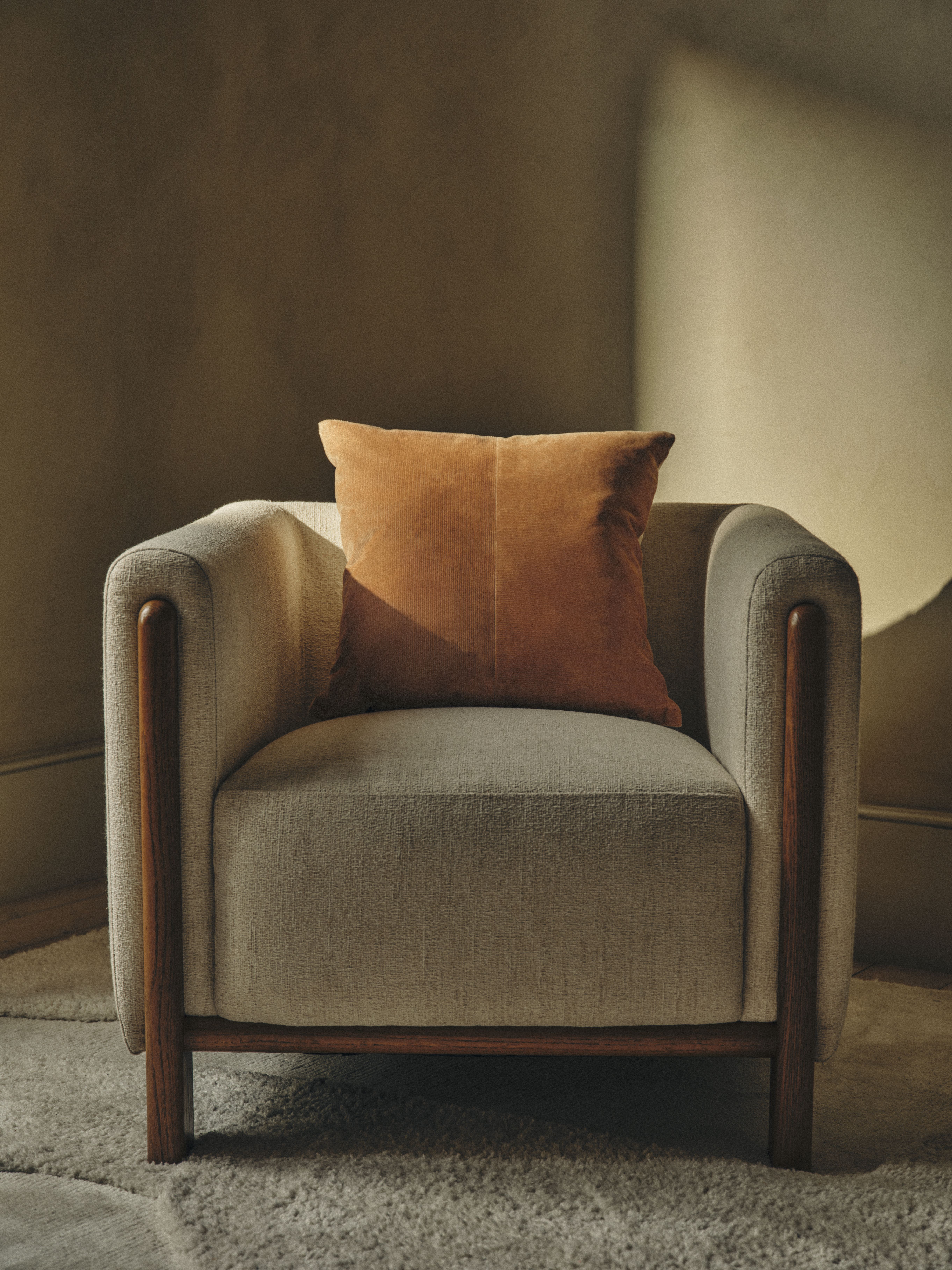 A light gray upholstered armchair with a dark wood frame and an orange corduroy cushion sits on a shaggy gray rug.