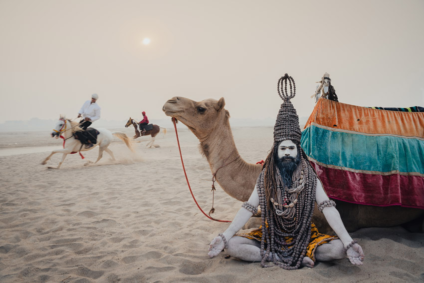 Man with long dreadlocks and ash-covered skin sitting on sand next to a camel, with men on horseback in the background.