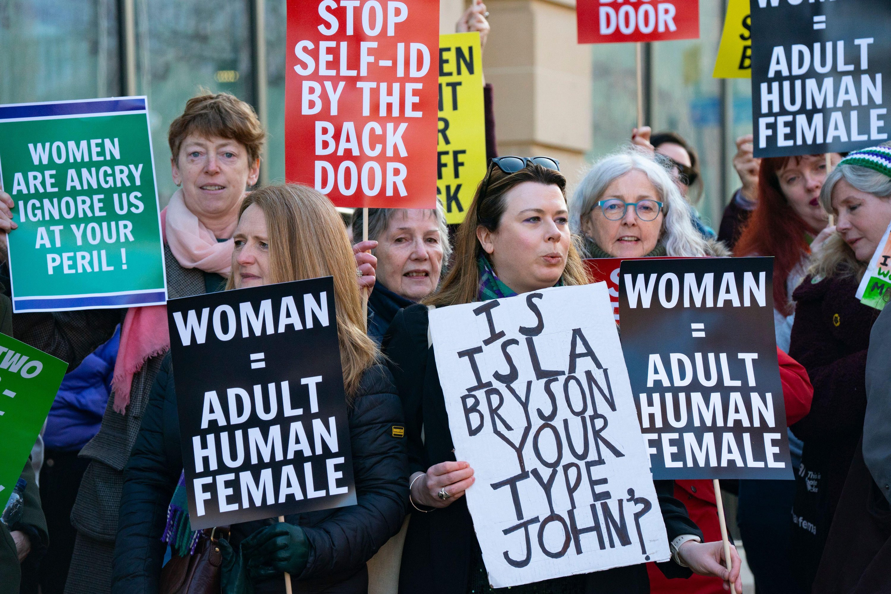 Women protest outside an International Women's Day event with signs reading "WOMAN = ADULT HUMAN FEMALE" and "STOP SELF-ID BY THE BACK DOOR".