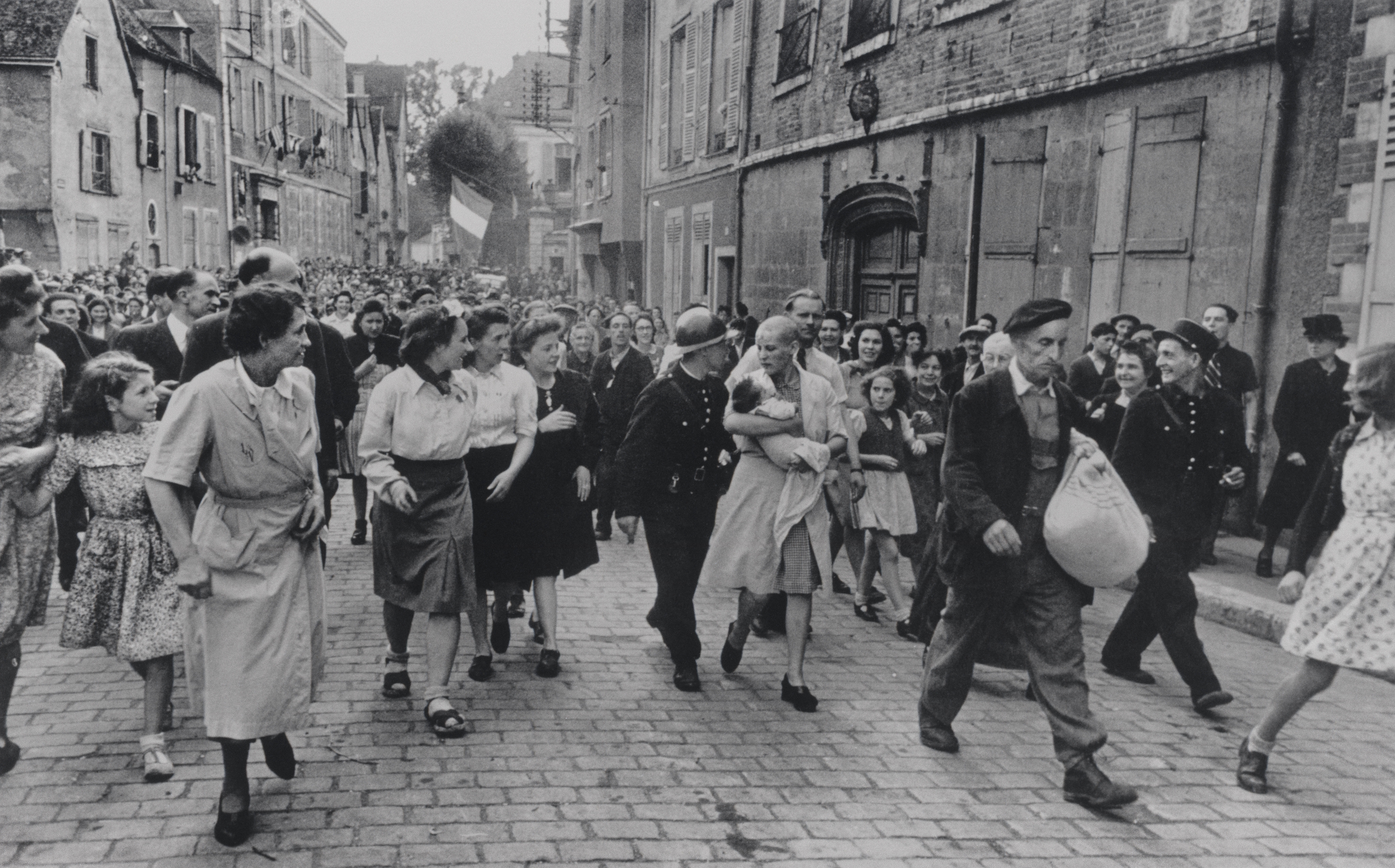 A shaved-head French woman carrying a baby is marched down a cobblestone street, surrounded by a crowd and a uniformed guard.