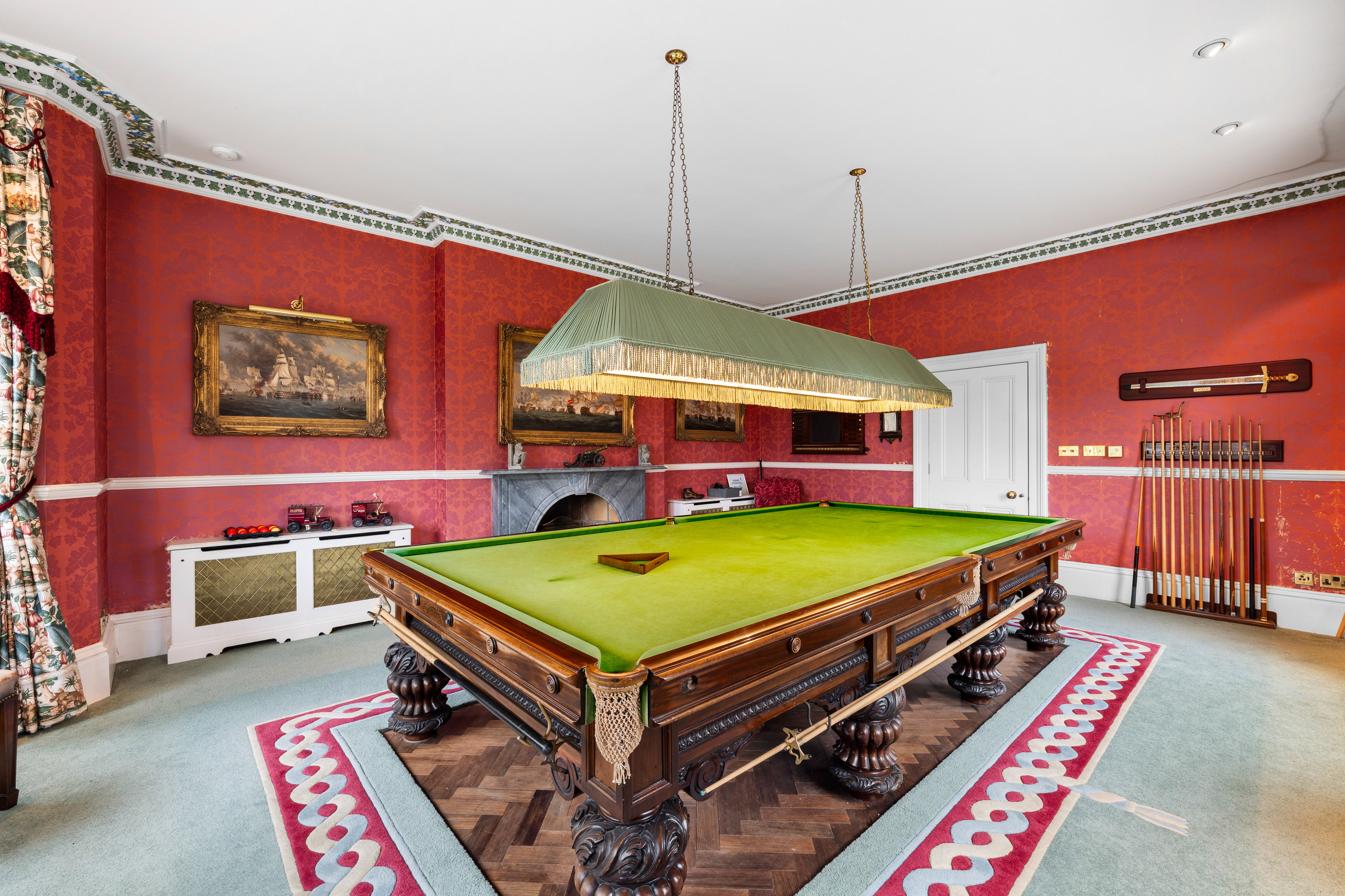 A billiards room with red patterned wallpaper, a green billiards table, and framed paintings.