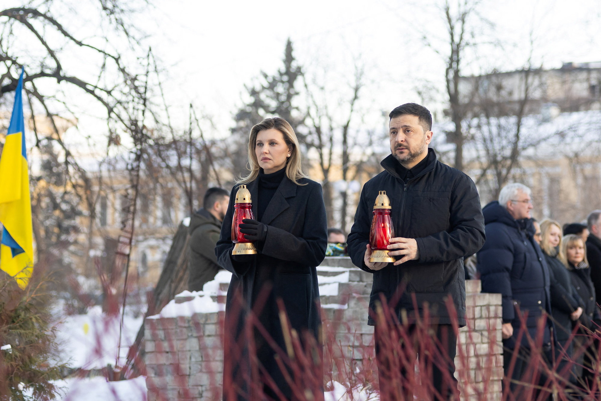 Volodymyr Zelenskiy and Olena Zelenska holding red memorial lamps at a monument.