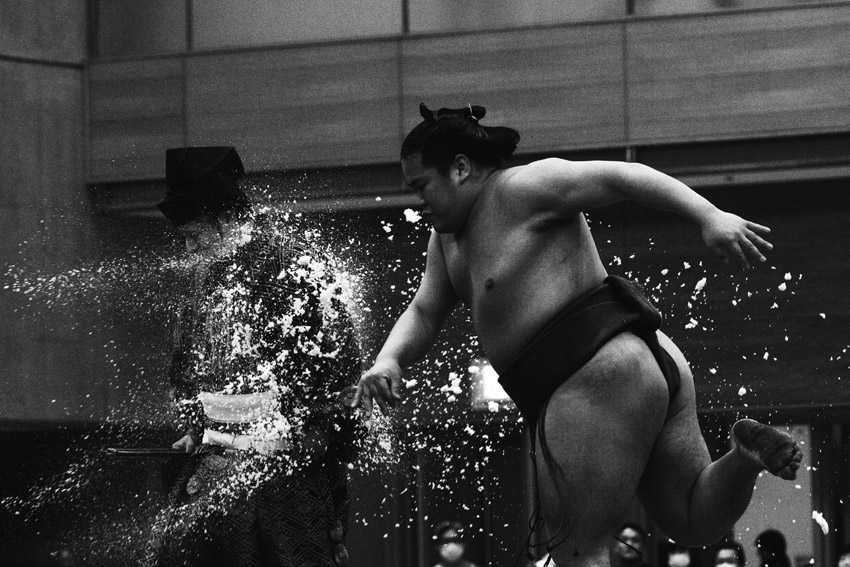 Black and white photo of a sumo wrestler throwing salt with a person dressed in traditional attire beside them.