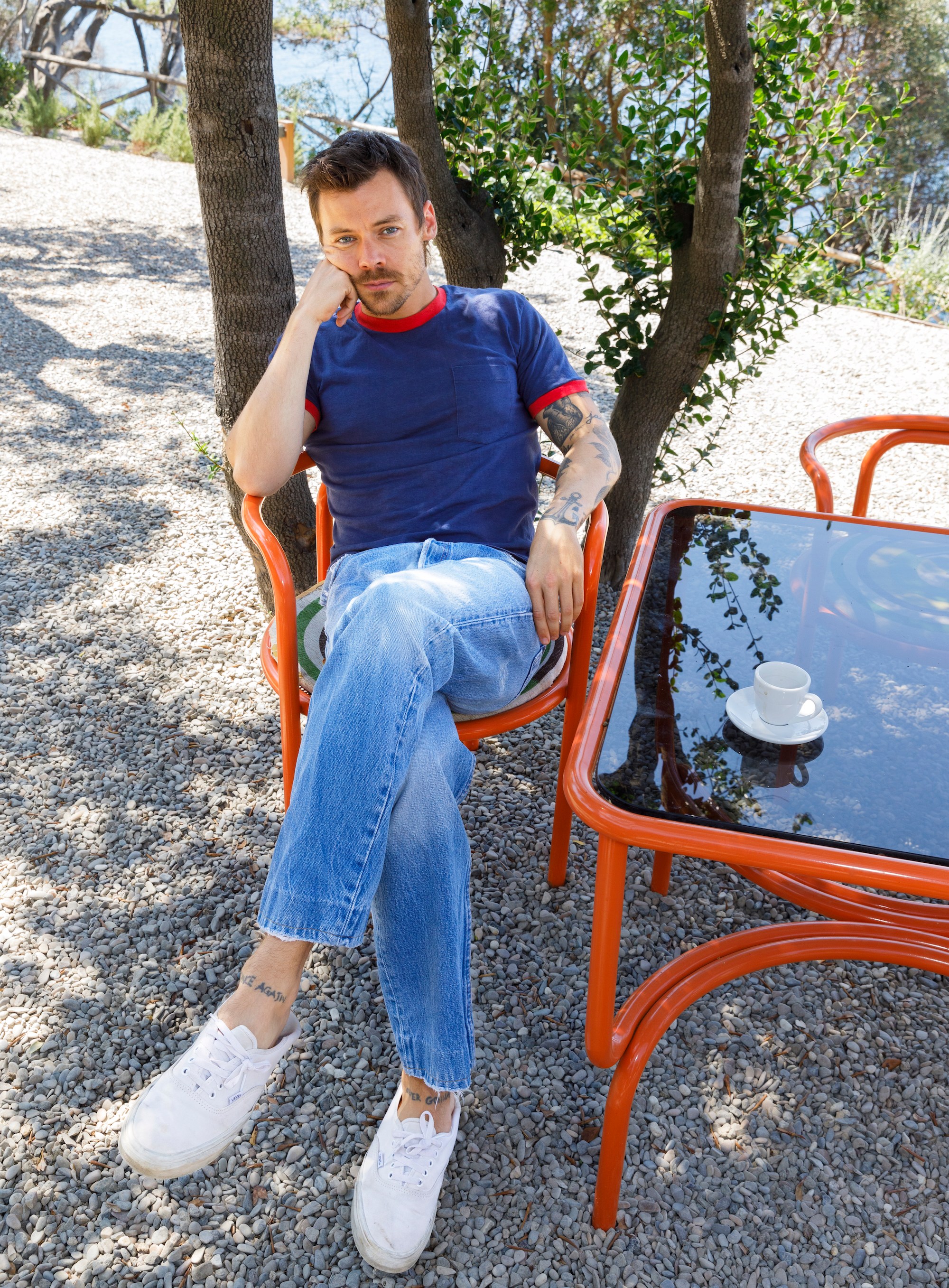 A man with a beard and tattoos sits in an orange chair under a tree, next to a table with a white teacup on it.