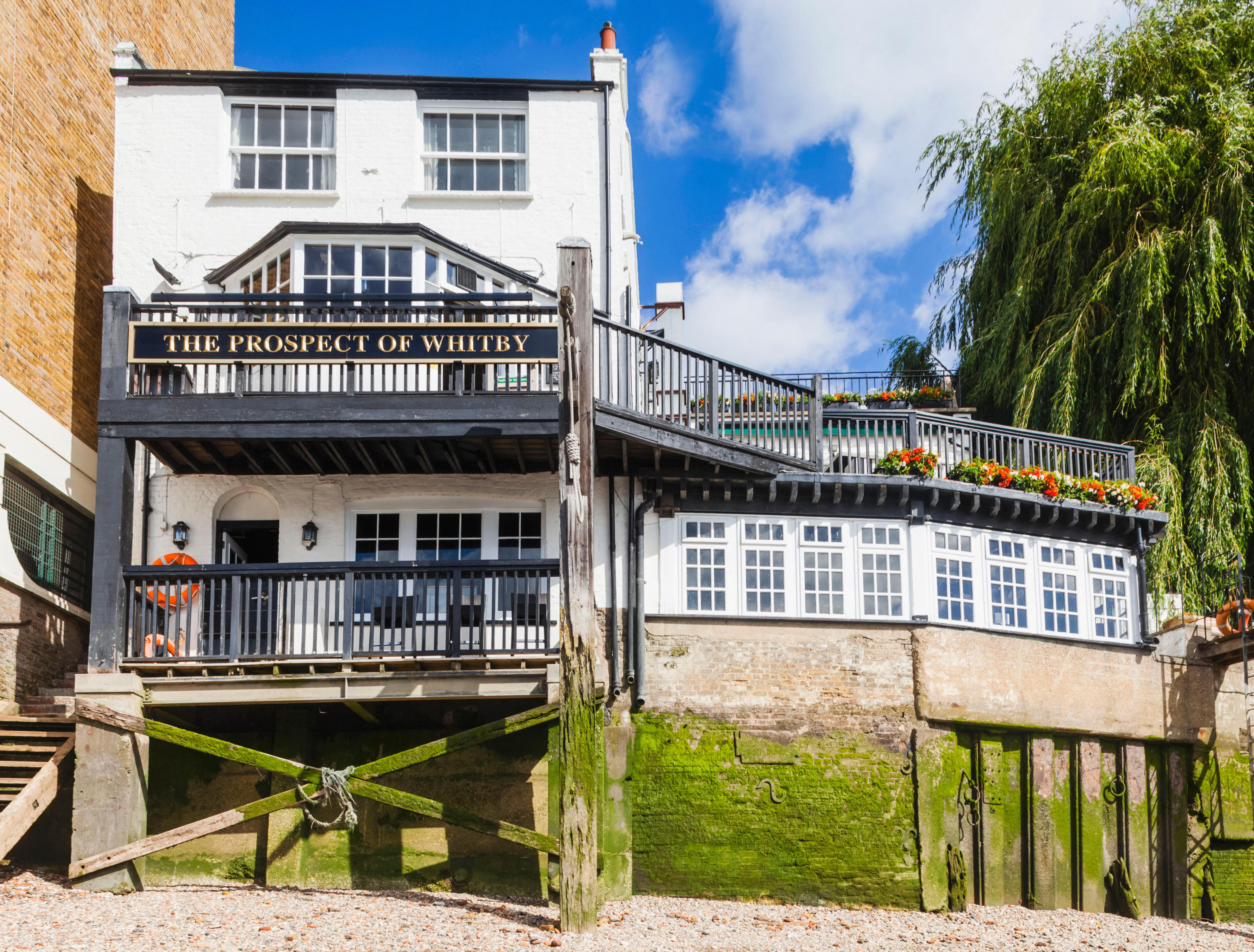 The historic riverside pub, The Prospect of Whitby, with its whitewashed walls, black railings, and flower boxes, sits on the bank of the Thames, with a wooden staircase leading up from the shingled shore.