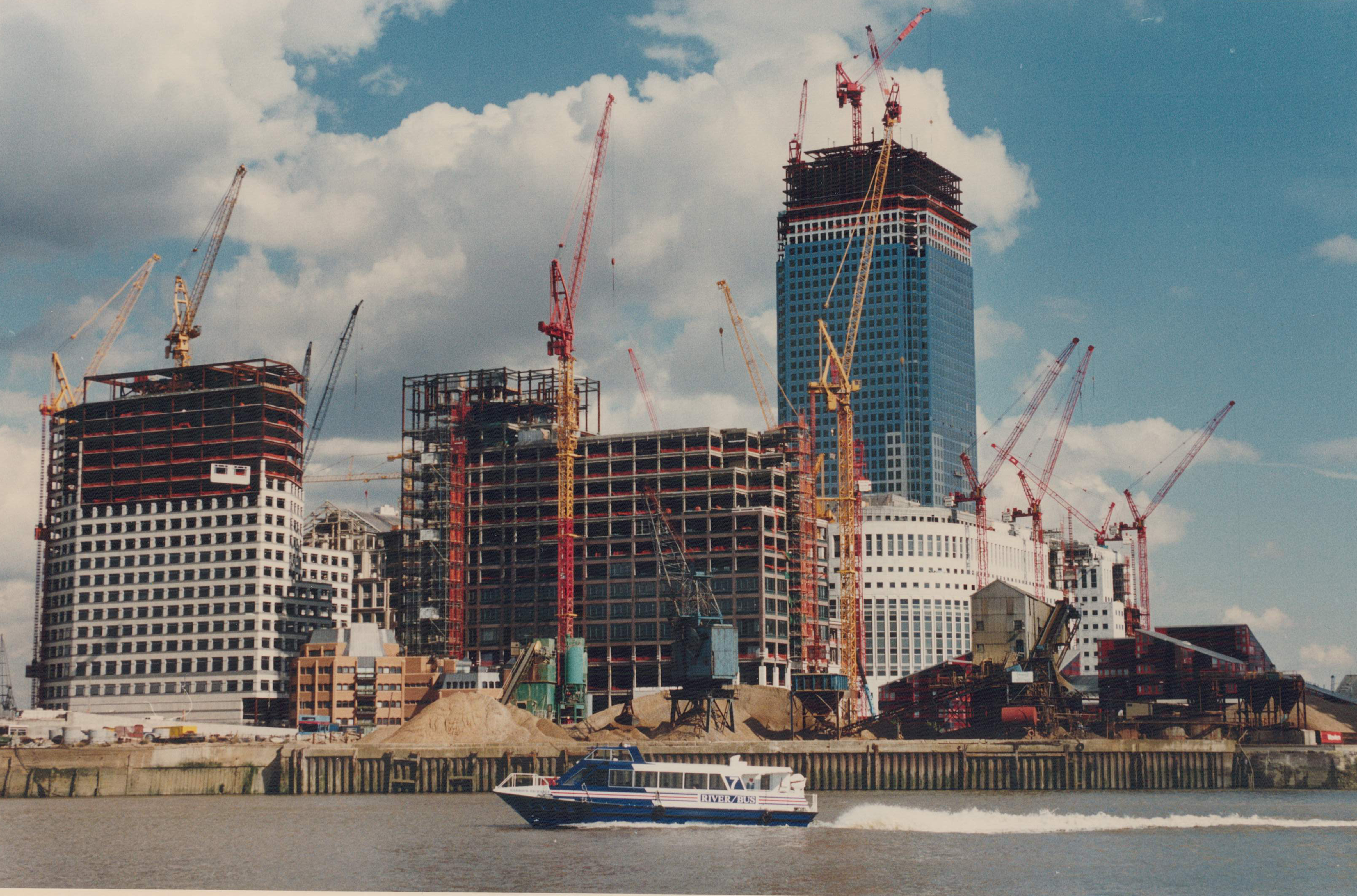 Number 1 Canada Square under construction, with surrounding buildings and cranes, viewed from the water with a River Bus in the foreground.