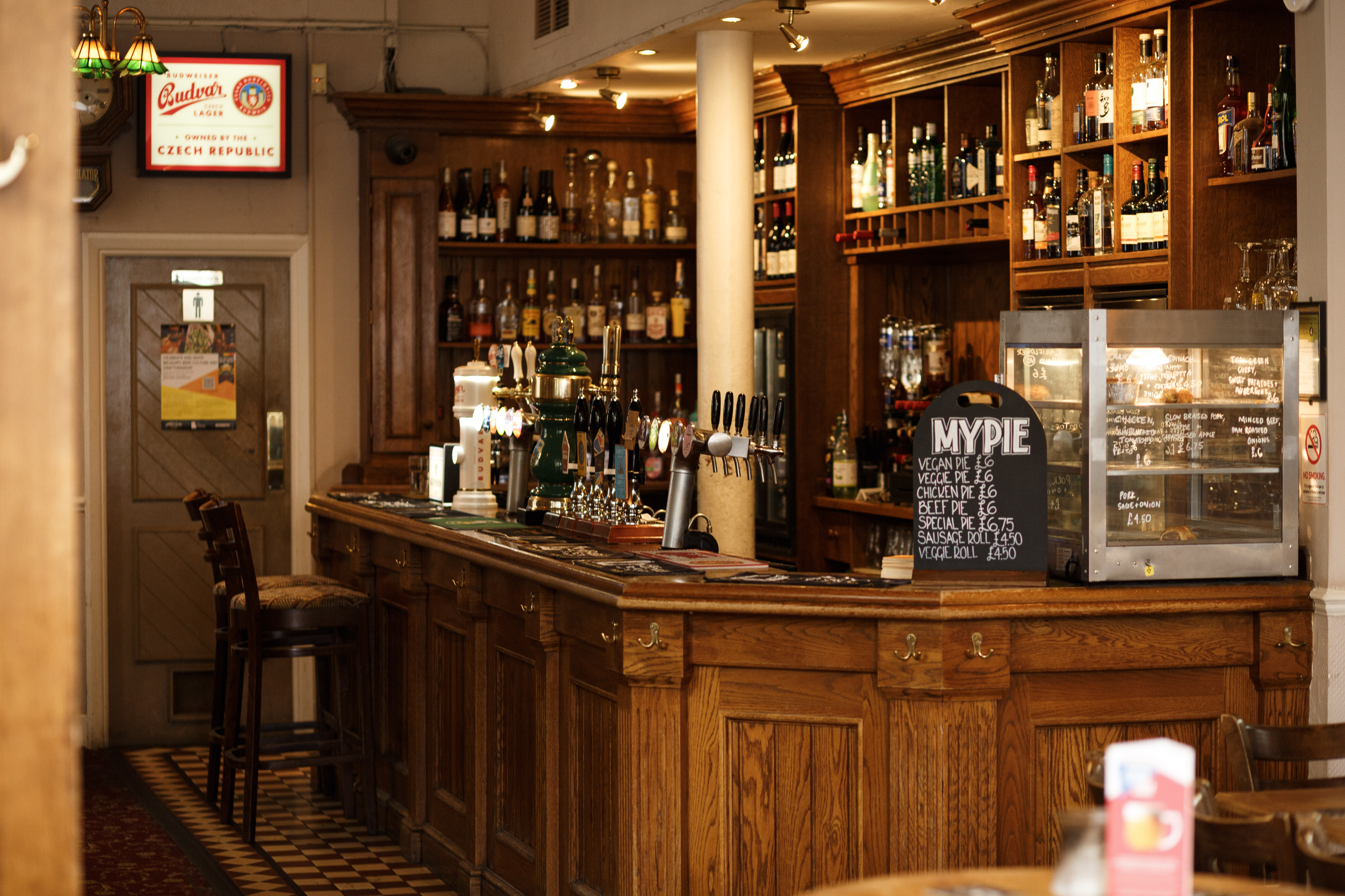 Interior of The Sutton Arms pub, showing the bar with beer taps and bottles of alcohol on shelves, and a menu board for "MyPie".