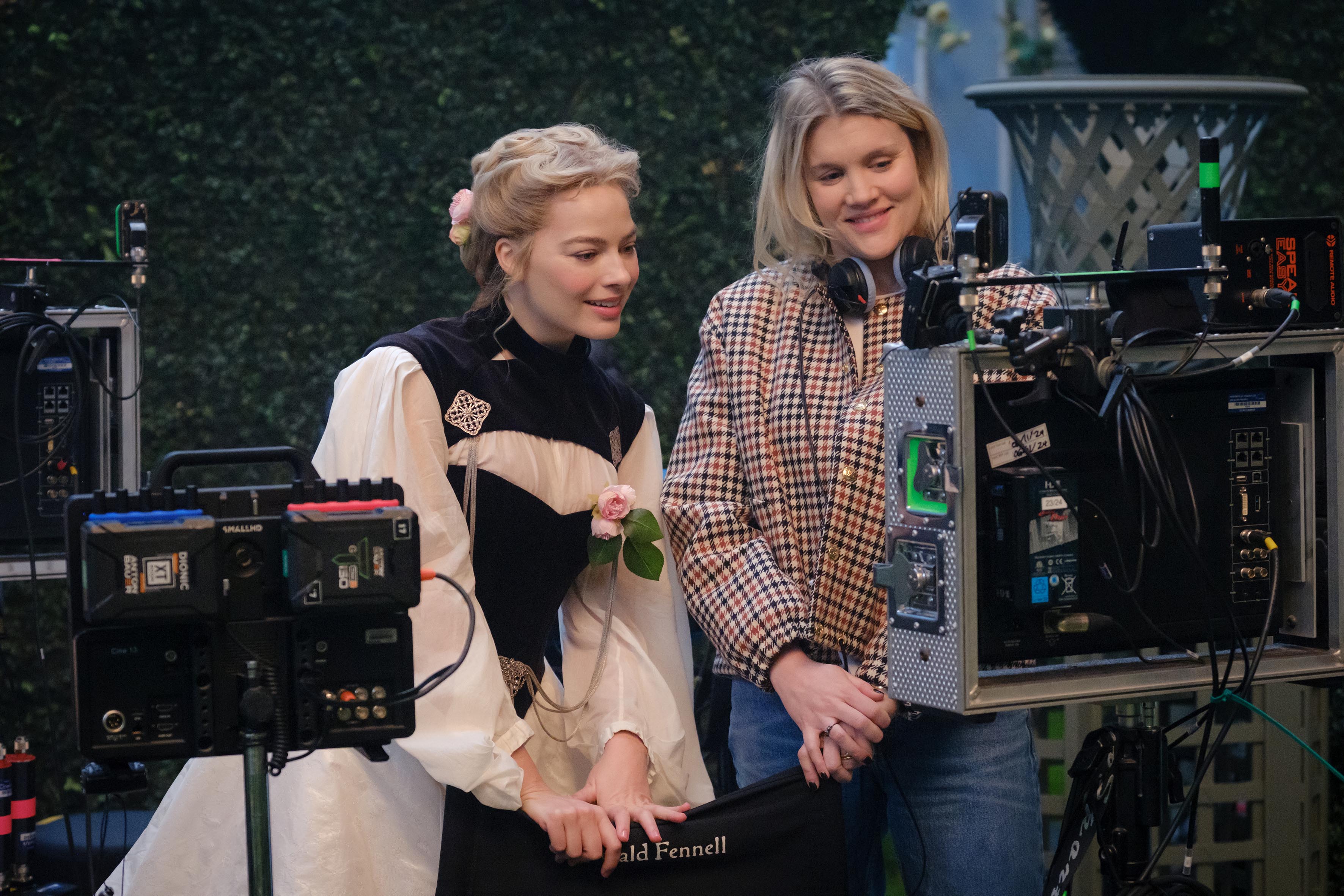 Margot Robbie, in period costume, and Emerald Fennell, in a checkered jacket, on a film set with camera equipment.