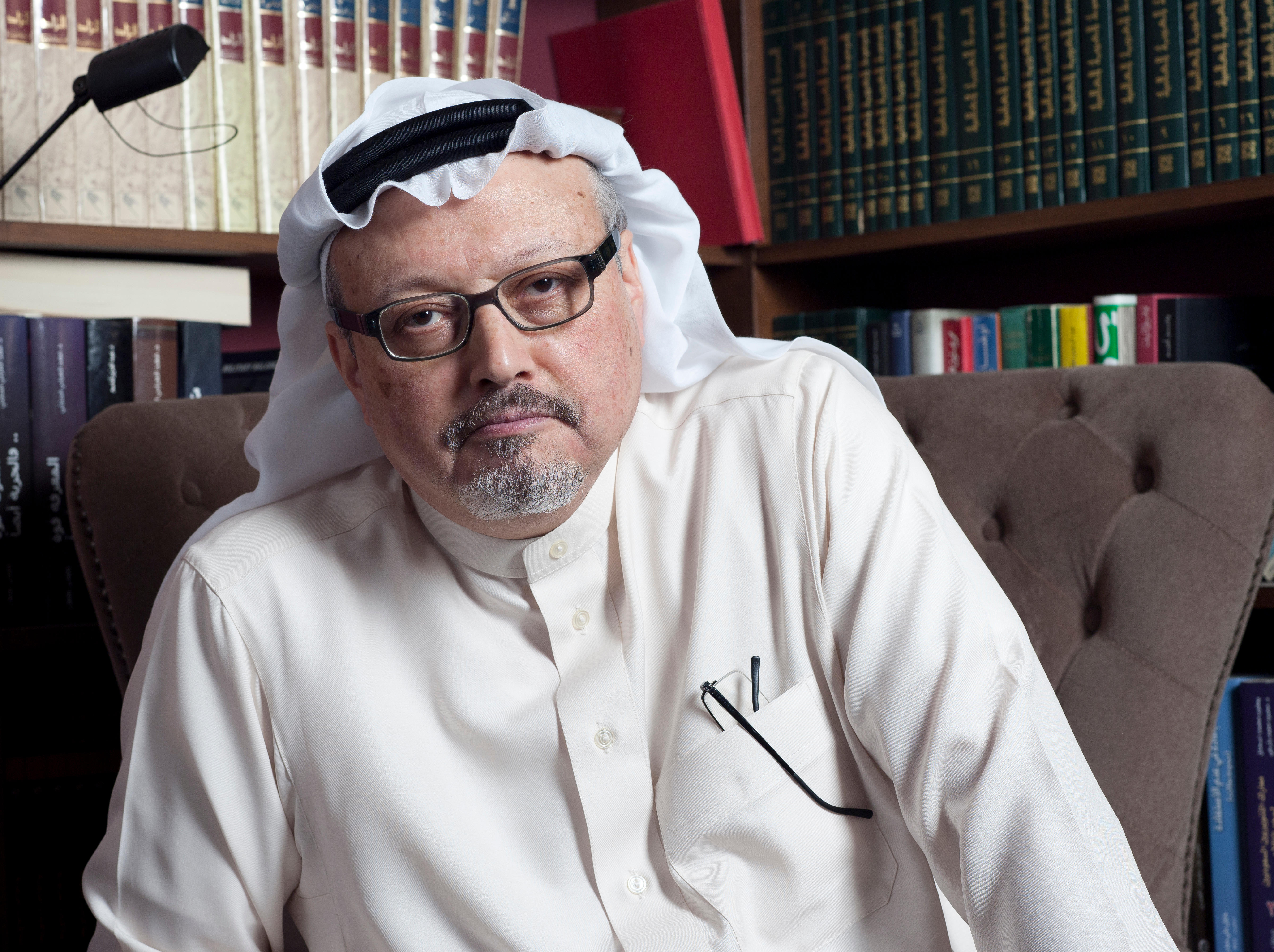 Saudi journalist Jamal Khashoggi sits in front of a bookshelf, wearing a white thobe and glasses.