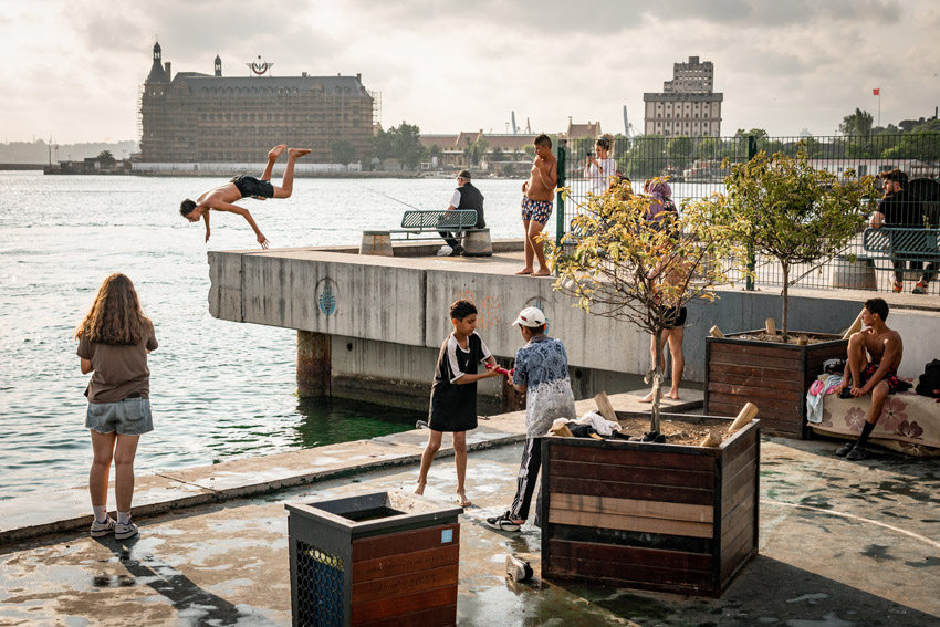A young man dives headfirst off a concrete pier into the water while other people relax, some fishing, and some kids play on the pier with a large building in the background.