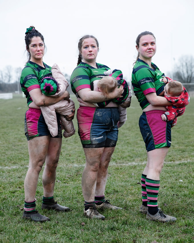 Three muddy women in rugby uniforms holding babies in a field.