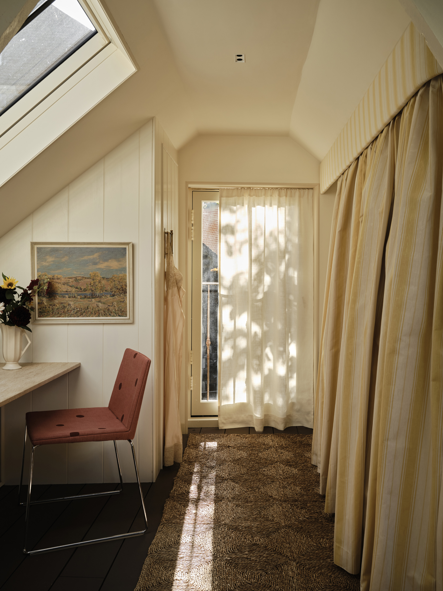 An attic room with a skylight, a red chair, a window with sheer curtains, and a long rug leading to an opening with striped curtains.