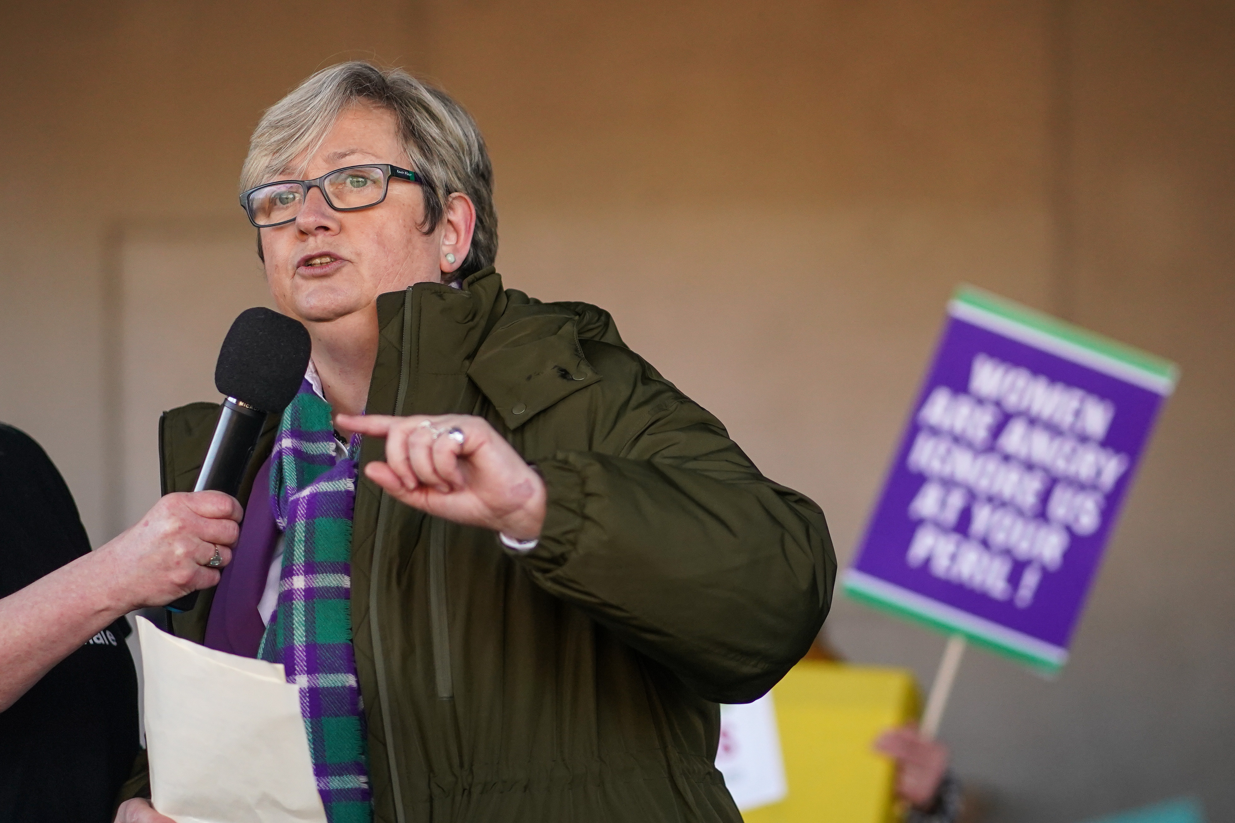 Joanna Cherry MP speaking into a microphone with a protest sign visible in the background.