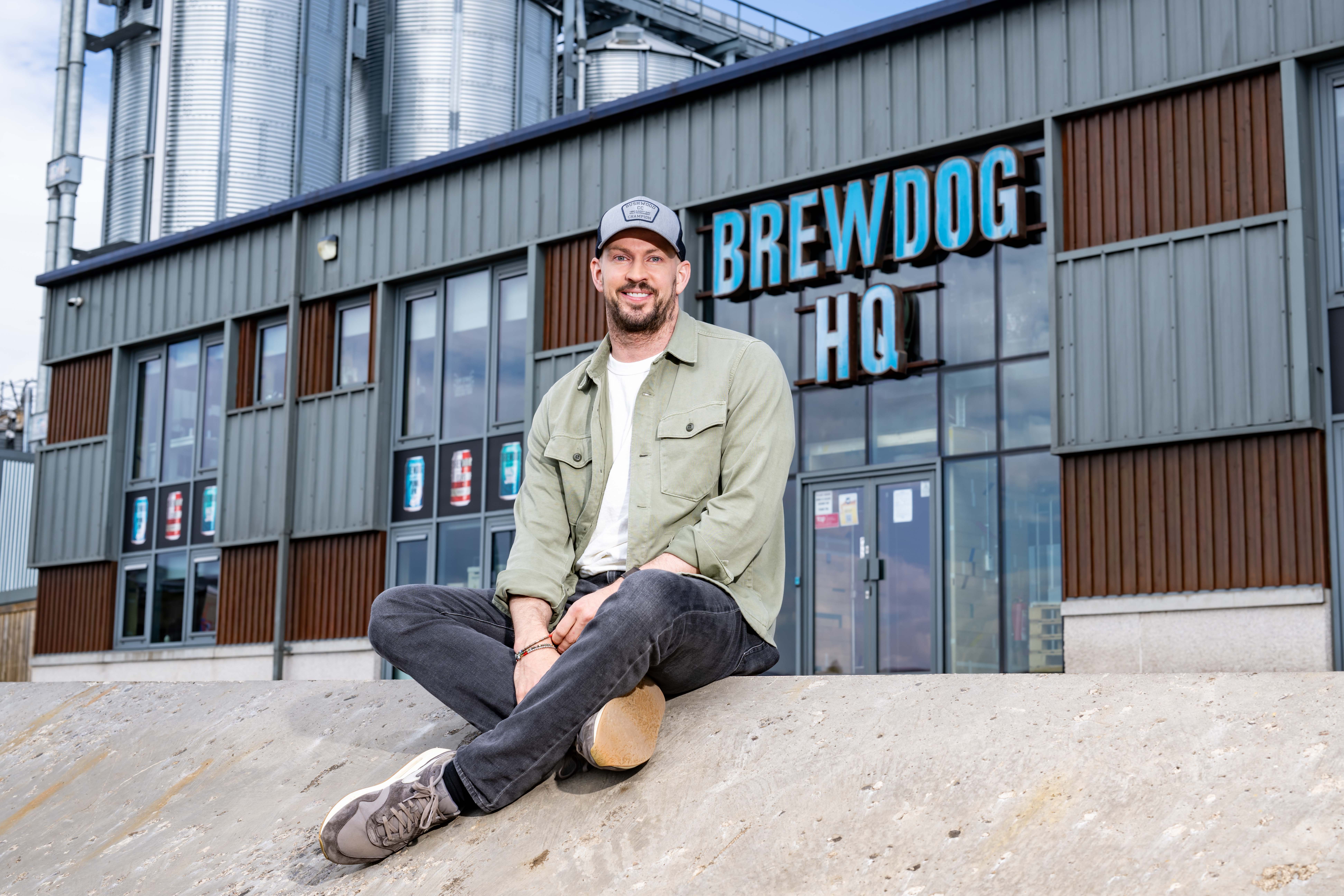BrewDog's James Watt smiling while seated in front of the BrewDog HQ building and large grain silos.