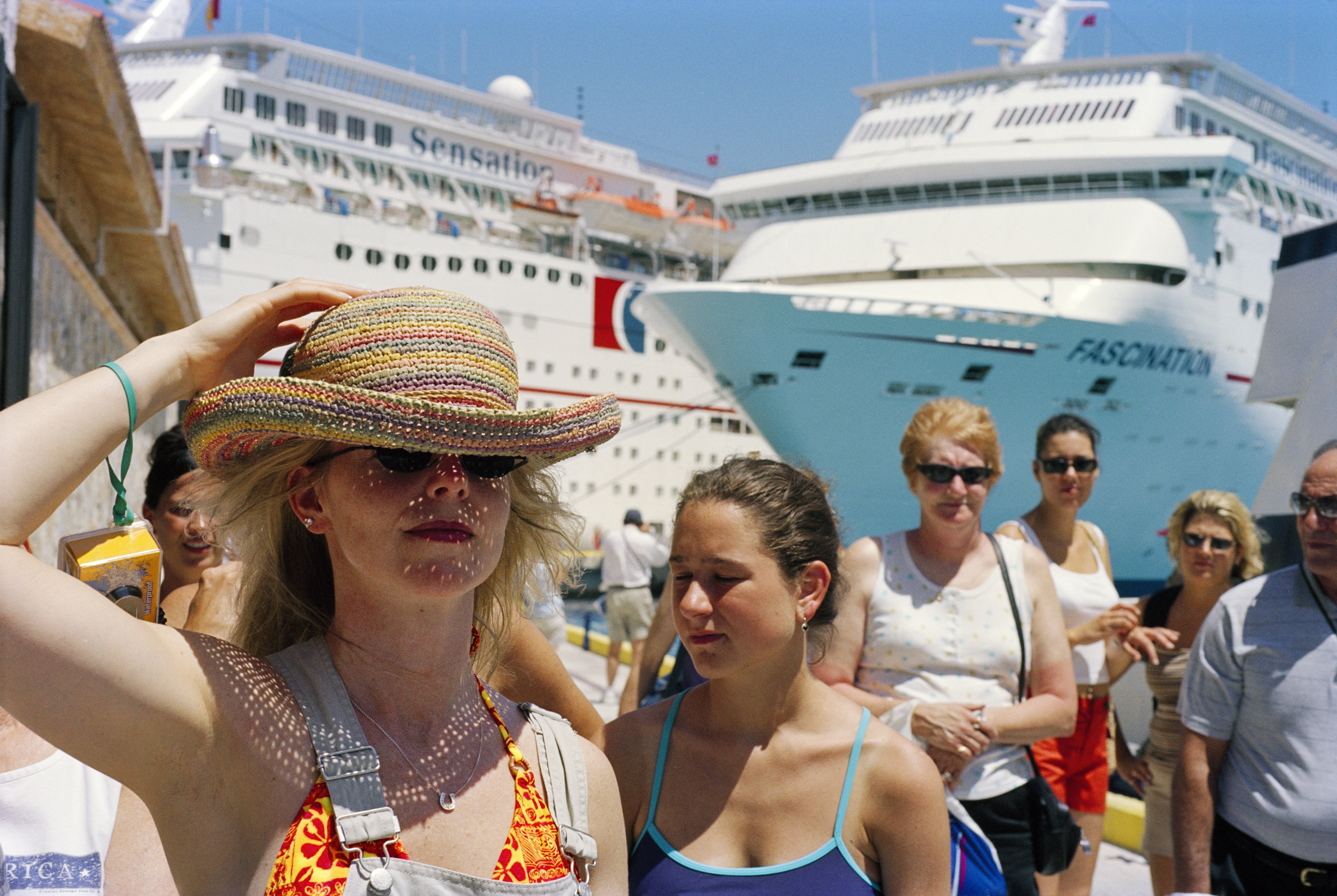 A woman in a straw hat and sunglasses stands in the foreground, with several people and two large cruise ships in the background.