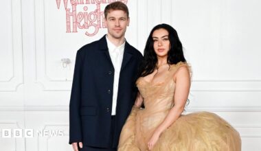 Finn Keane and Charli XCX standing next to each other at the Wuthering Heights premiere. There is a white wall behind them. Finn is wearing a white shirt with a dark navy jacket unbuttoned and is staring directly at the camera. Charli has her long black hair down and is wearing a gold dress.