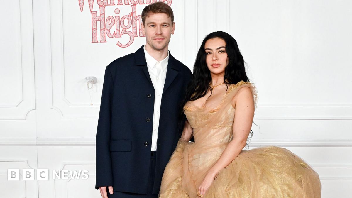 Finn Keane and Charli XCX standing next to each other at the Wuthering Heights premiere. There is a white wall behind them. Finn is wearing a white shirt with a dark navy jacket unbuttoned and is staring directly at the camera. Charli has her long black hair down and is wearing a gold dress.